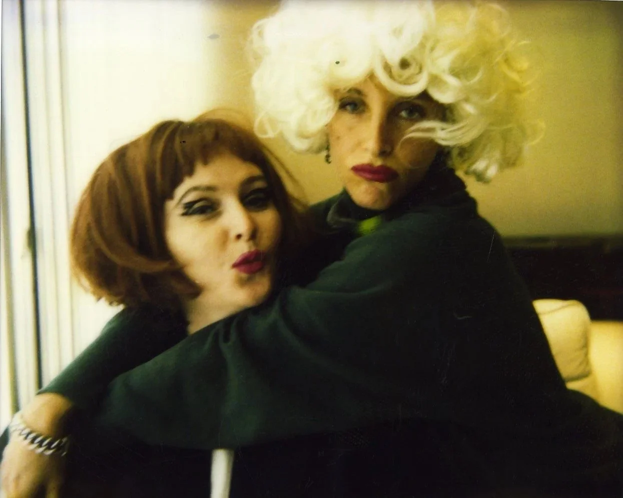 Two women dressed in drag with elaborate makeup and wigs posing closely together indoors.
