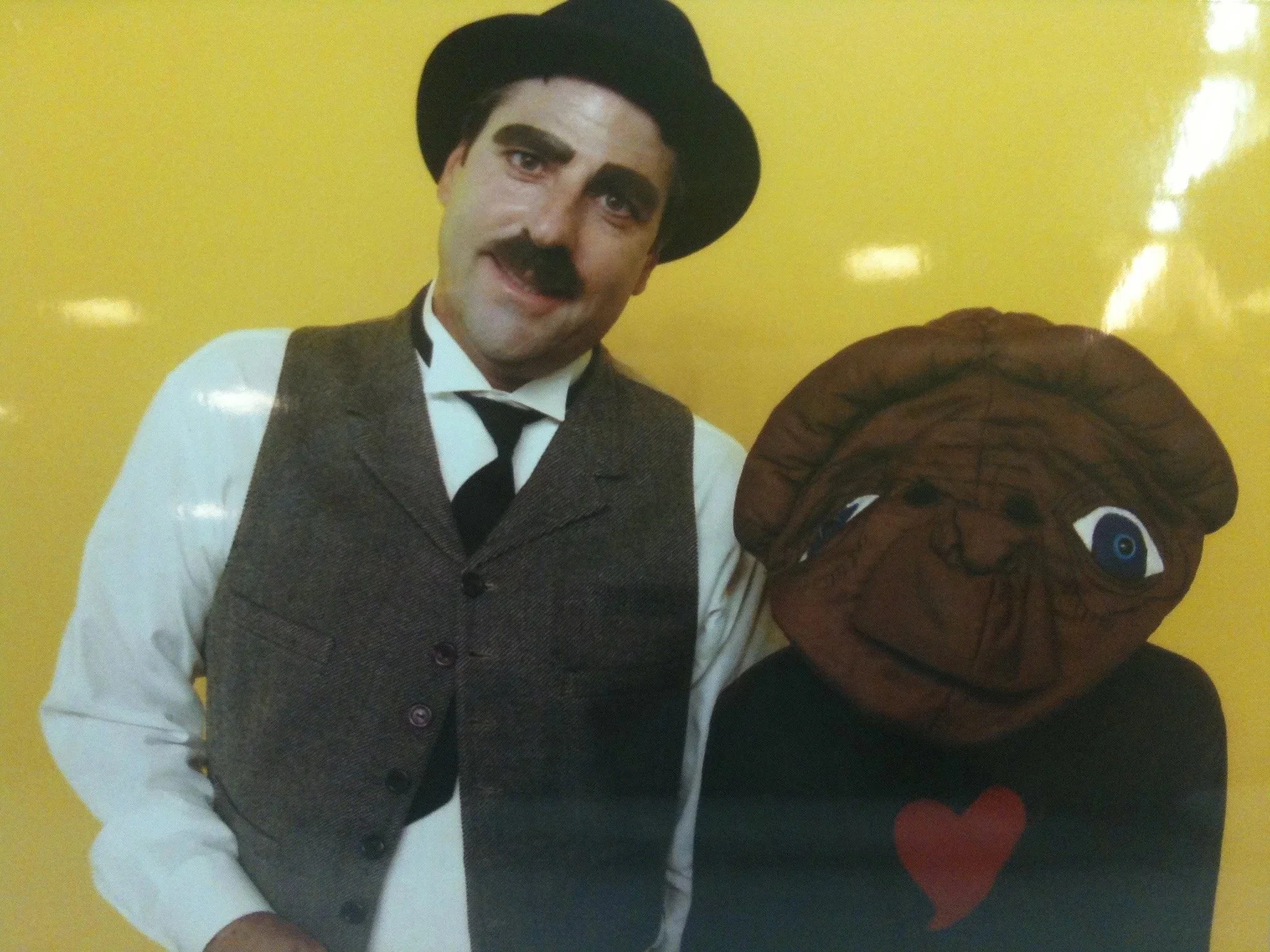 A man dressed in vintage clothing, including a vest and bow tie, standing against a yellow background with a mascot of a brown puppy and three red hearts on the right side.