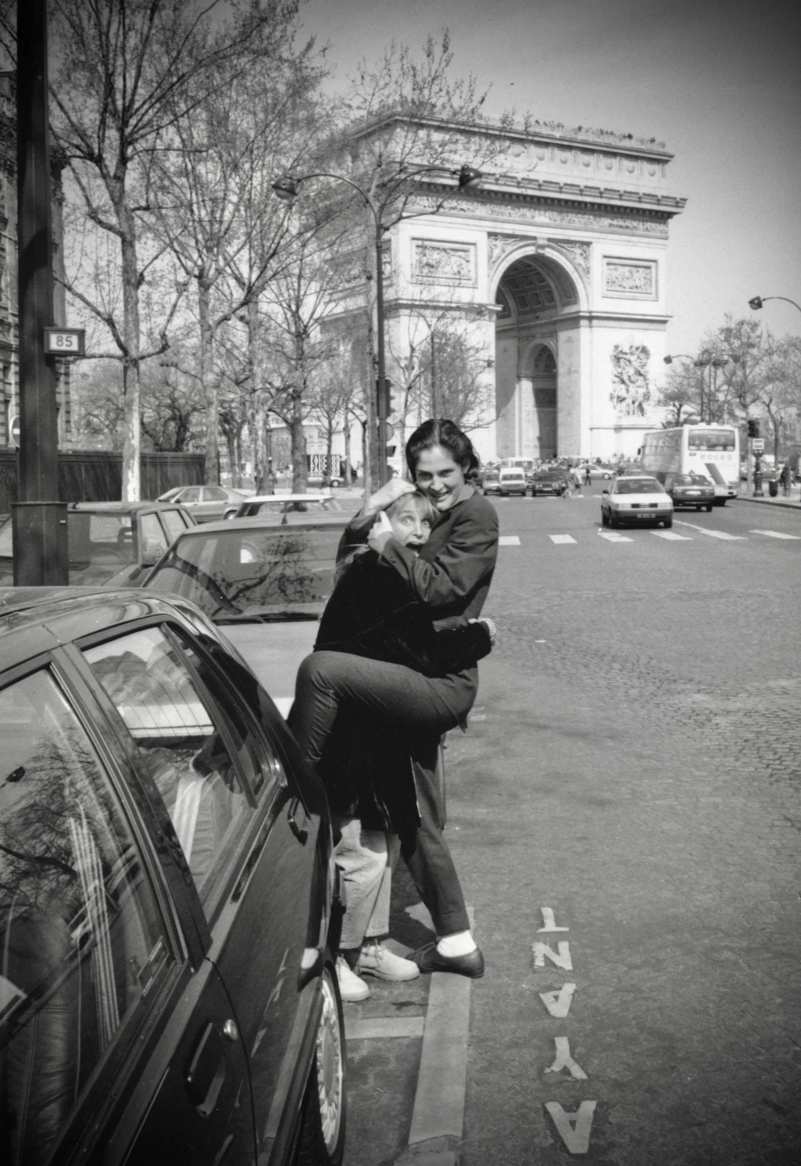 Two women, one with short blond hair and the other with dark curly hair, embracing on a Paris street in front of a car, with the Arc de Triomphe in the background.