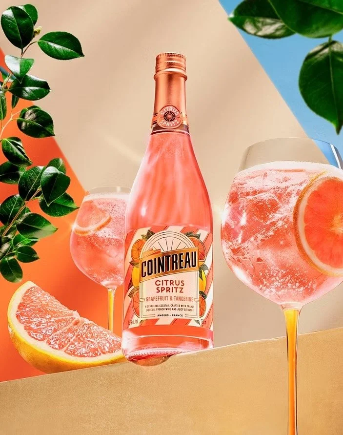 A bottle of Cointreau Citrus Spritz with grapefruit and tangerine flavor is placed on a surface alongside two large glass filled with grapefruit-infused sparkling drink, garnished with slices of grapefruit. Green leaves and colorful background add to