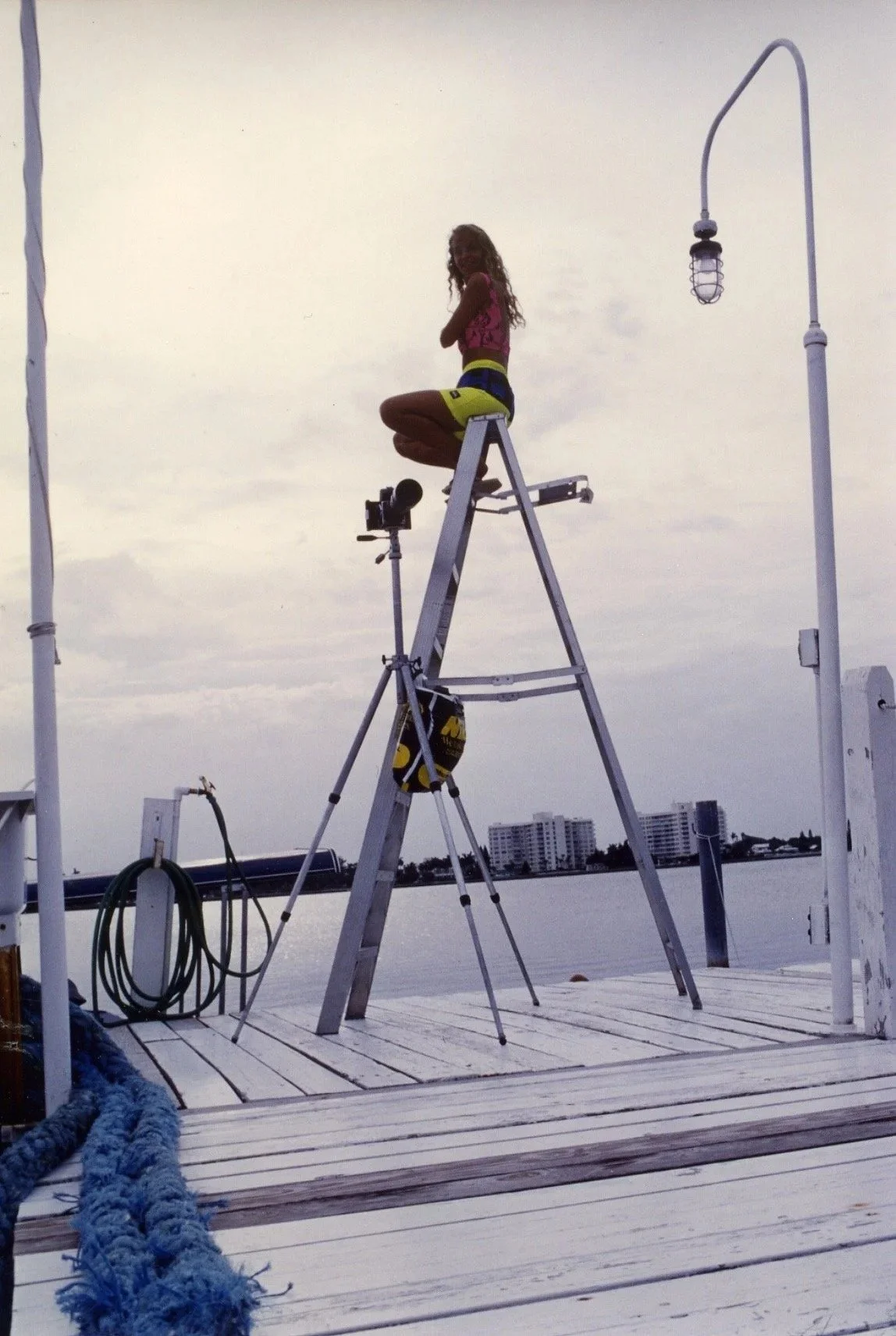 A person sitting on top of a tall ladder on a dock by the water, with city buildings in the background, lit by a hanging outdoor light, during overcast weather.