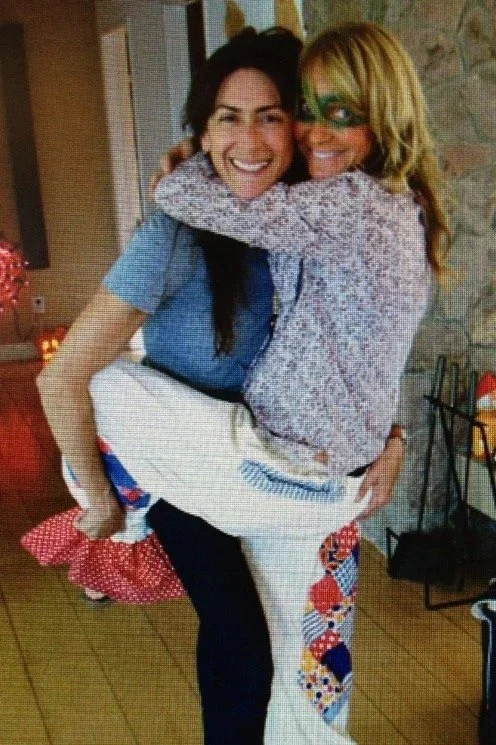 Two women hugging and smiling, one wearing a blue shirt and the other wearing a sweater with a green eye mask, in a cozy indoor setting.