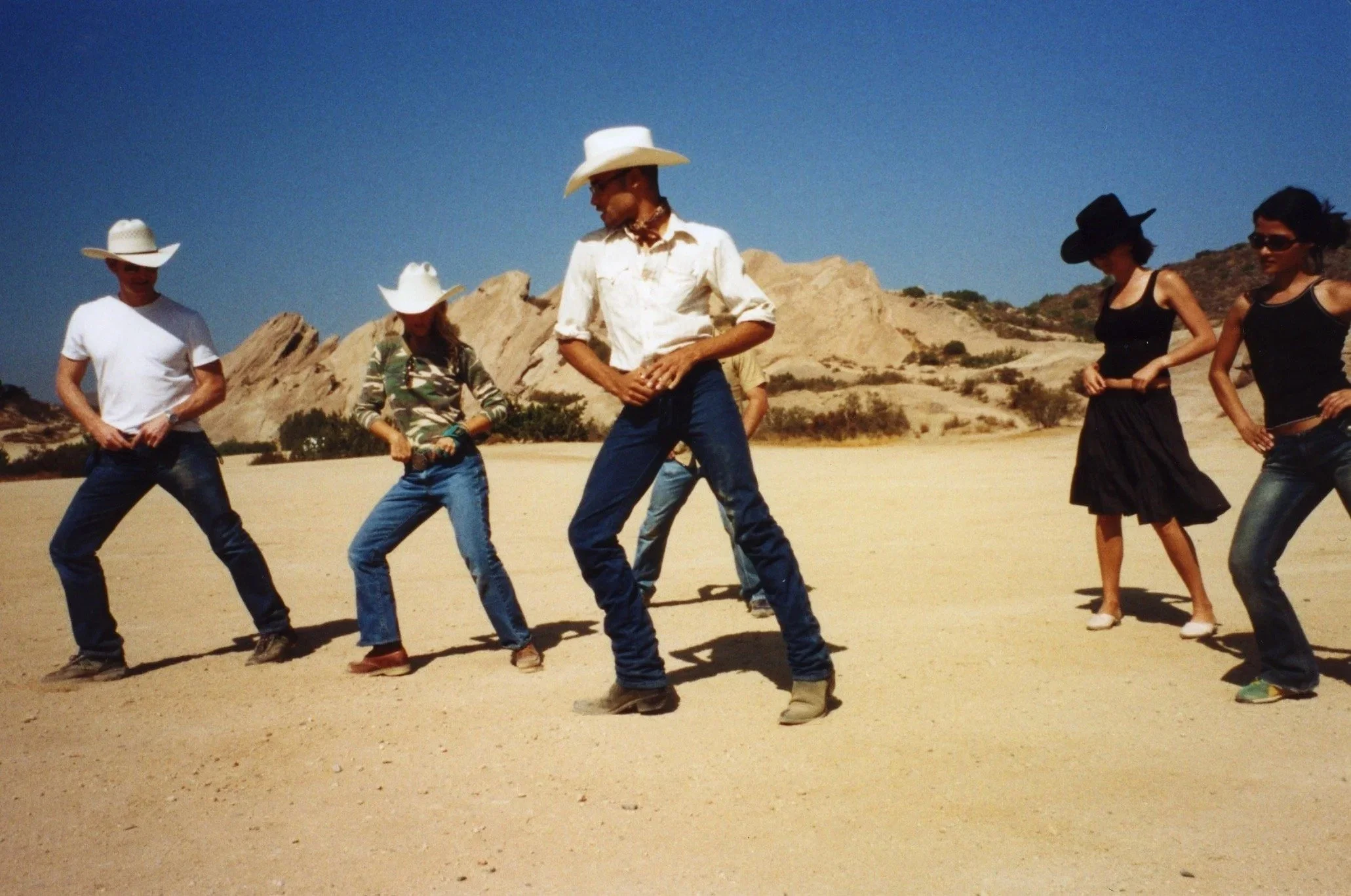 A group of people dressed in Western-style clothing, standing and posing on a desert landscape with rocky hills in the background, under a clear blue sky.