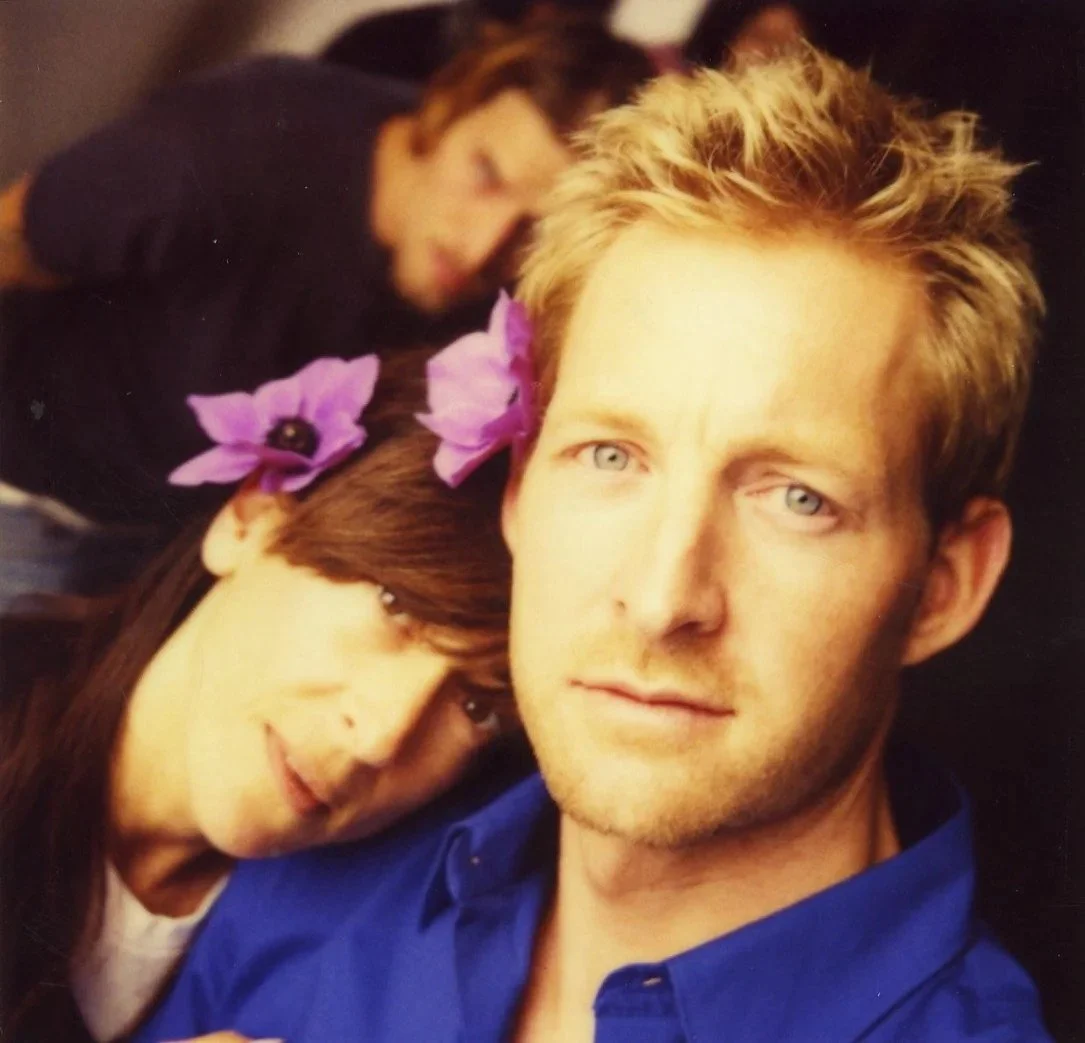 A woman and a man are close together, with the woman resting her head on the man's shoulder. The woman has long brown hair with purple flowers tucked behind her ear. The man has short blonde hair and blue eyes, wearing a blue shirt.