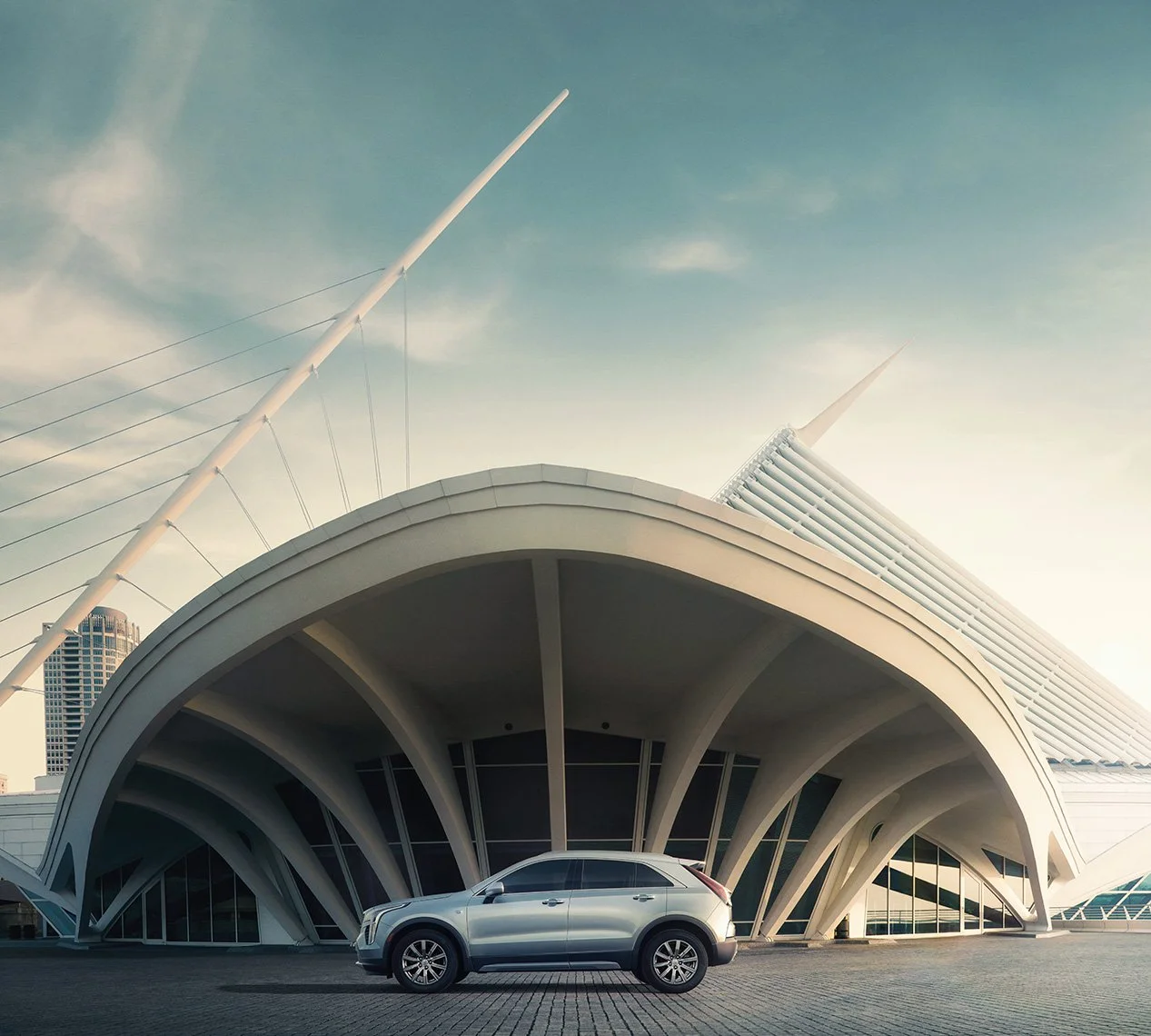 A modern architectural building with a curved white facade and large glass windows, featuring tall structural elements and strings of cables extending upward, with a silver SUV parked in front.