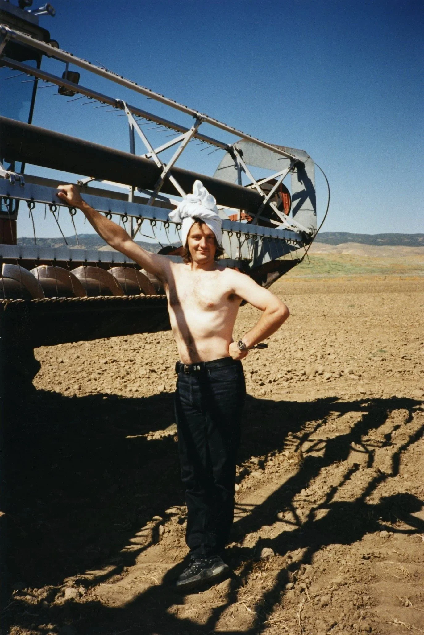 A shirtless man with a white cloth wrapped around his head, standing on dirt ground in front of large farming equipment in a rural landscape with hills in the background.