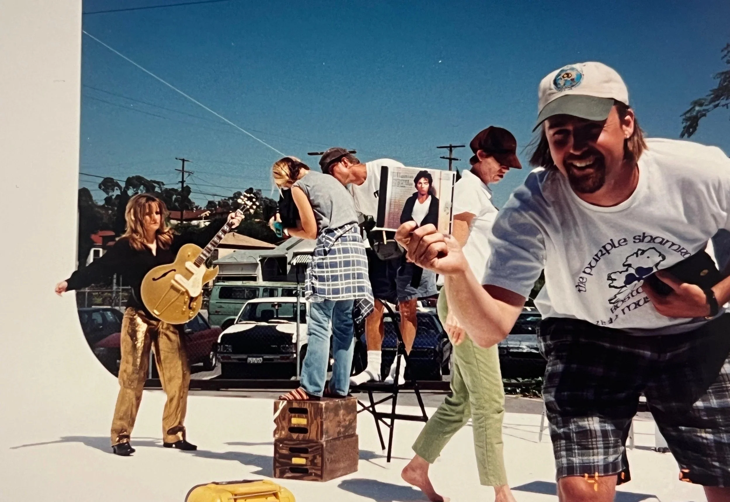 A group of people gathered outdoors on a sunny day, with one person playing a guitar, another holding a framed photograph, and others engaged in conversation or activities, with parked cars and utility poles in the background.