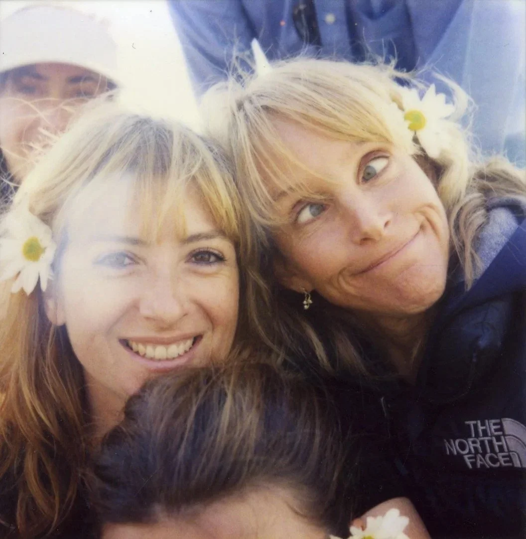 Four women taking a selfie together, two of them with daisies in their hair, smiling and making amusing faces.