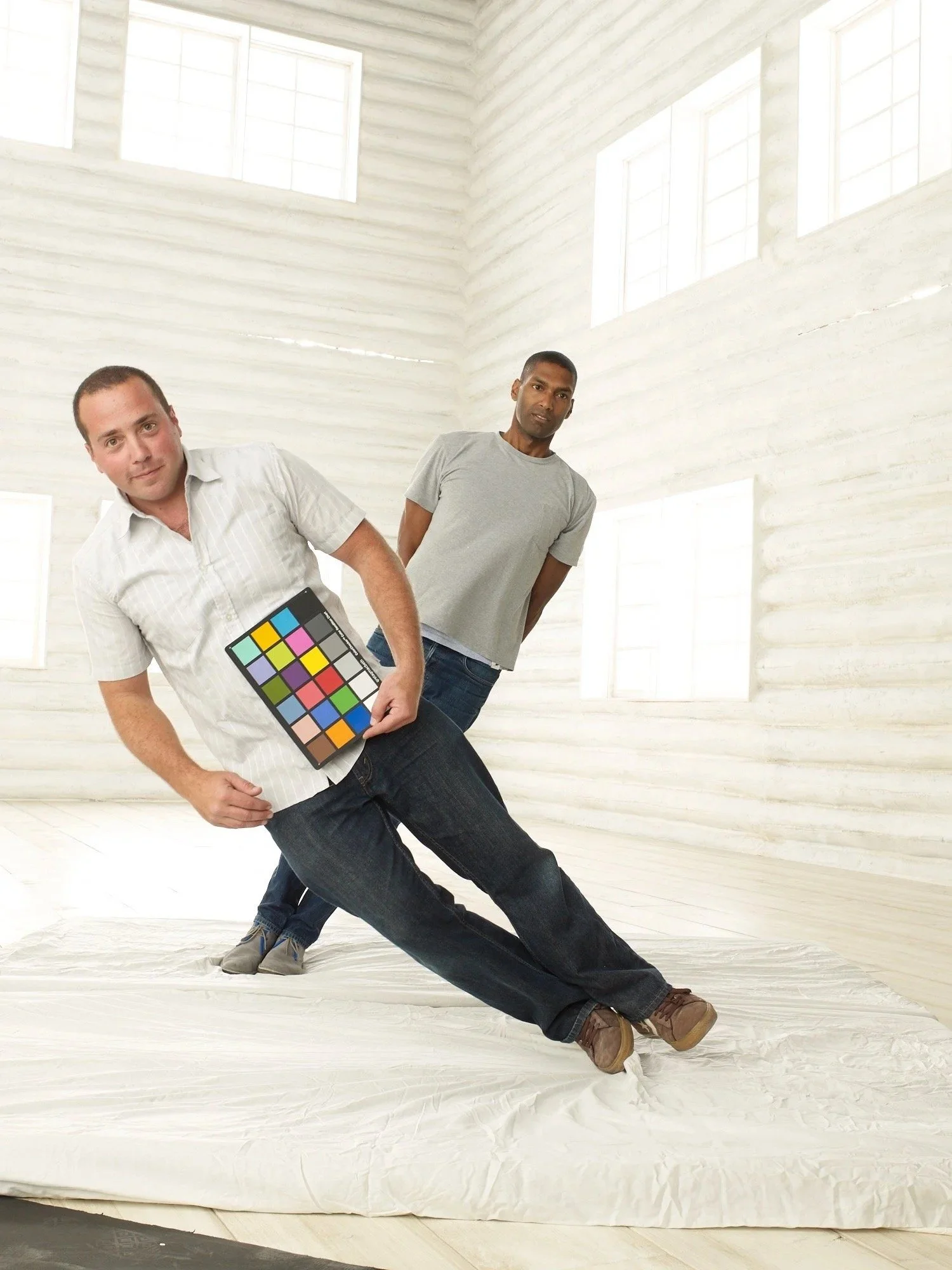 Two men in casual clothing standing in a bright, white room with large windows, with one holding a color calibration chart.