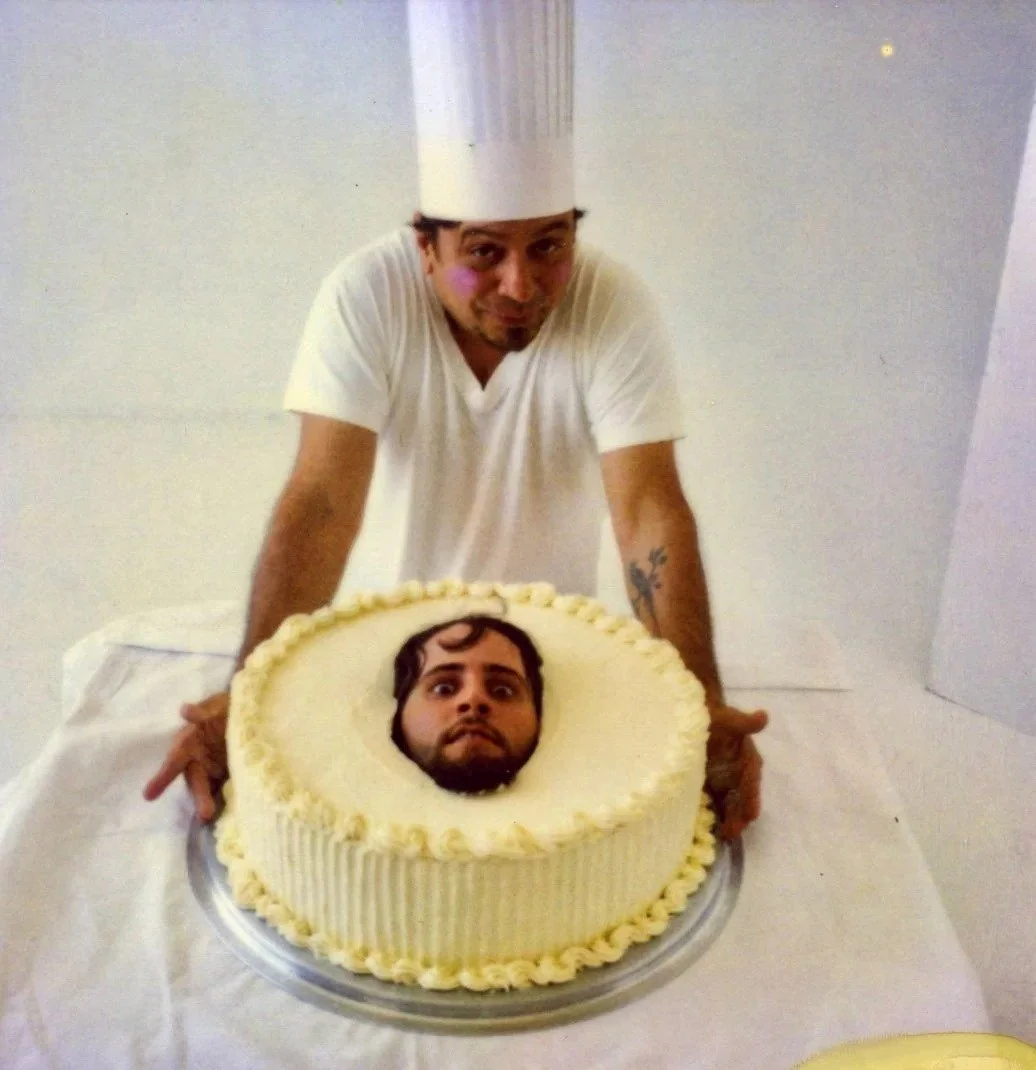 A chef wearing a tall white hat and white shirt stands behind a cake with a cut-out center. A man's face is visible through the hole in the cake, making it look like he has a large head on the cake.