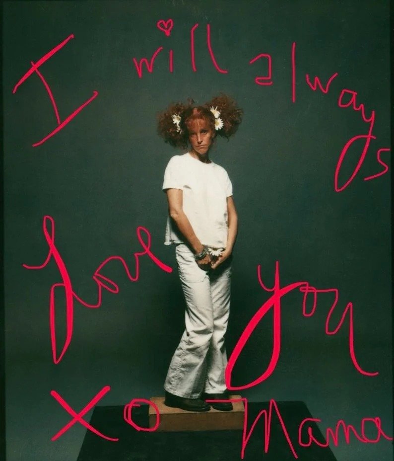 A girl with curly hair and white flowers in her hair standing on a small wooden platform against a dark green background. There is pink handwritten text around her that says, "I will always love you" and "Xo Mama."