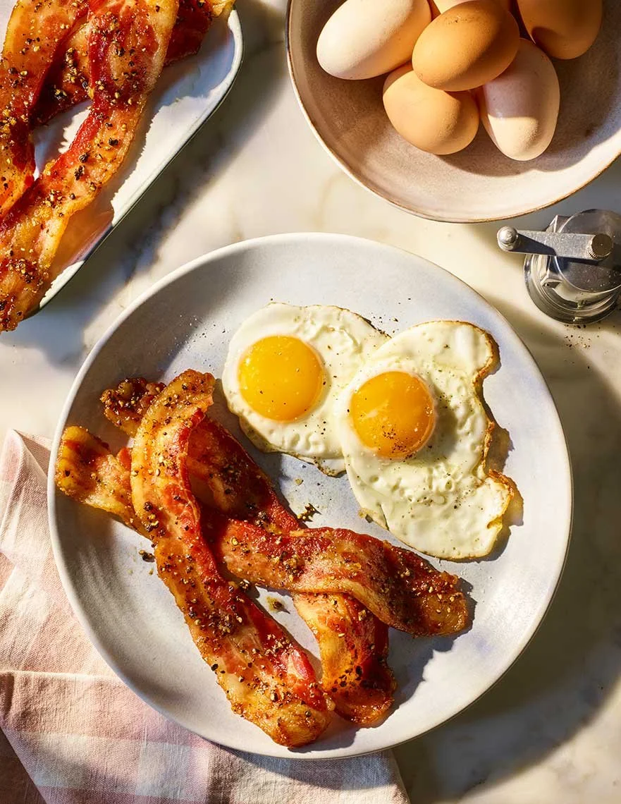 A breakfast plate with two sunny side up eggs and crispy bacon, accompanied by a bowl of eggs, a pepper grinder, and a side of baked bacon strips.