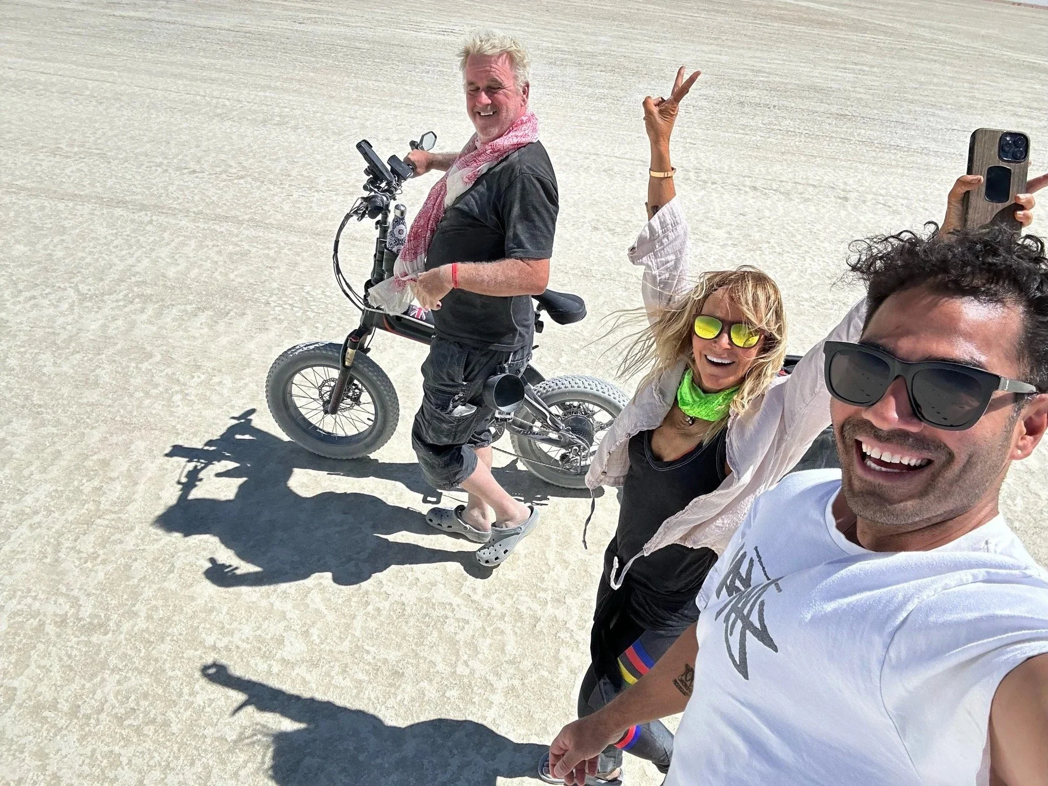 A group of people taking a selfie on a white salt flat, with one person on a bike and others smiling and showing peace signs.