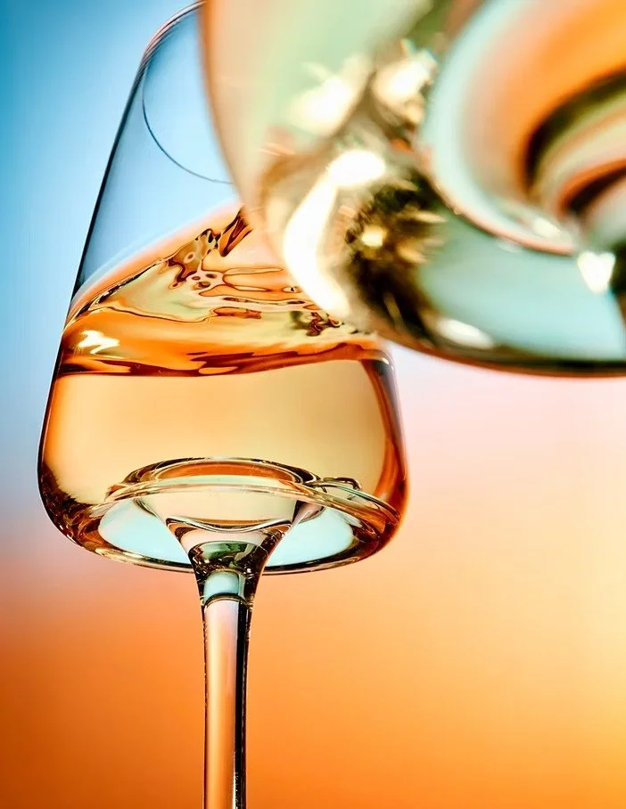 A close-up of a wine glass filled with rosé wine, with another glass pouring wine into it, against a colorful blurred background.