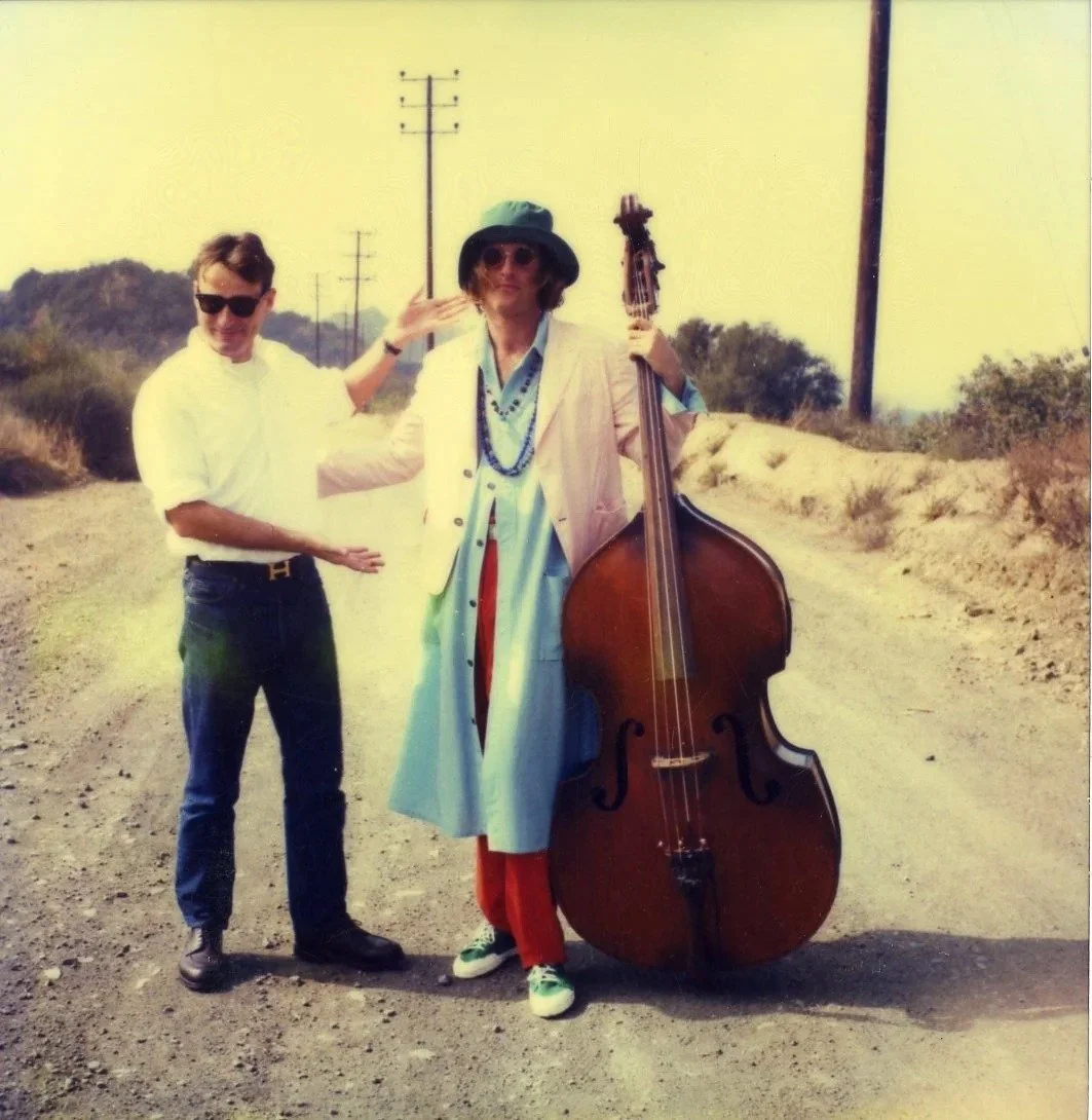 Two men standing on a dusty road, one holding a double bass and the other gesturing towards him, with power lines and distant hills in the background.