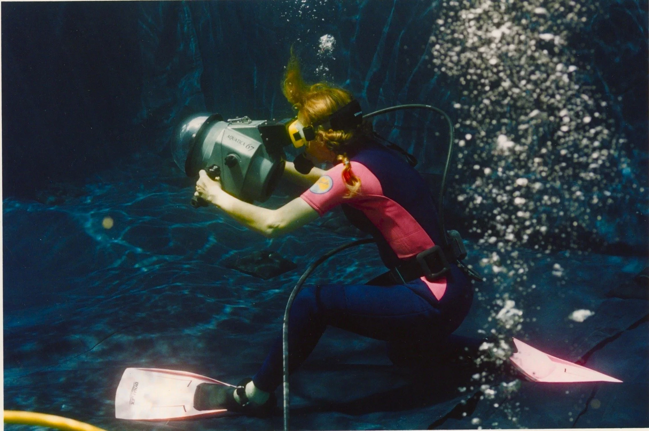 A person with long hair in a braid wearing a pink and black wetsuit underwater, operating a device labeled 'Aquatic 67' while observing marine life.