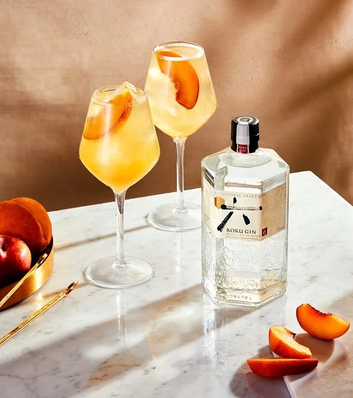 Two glasses of peach cocktail with ice and peach slices, a bottle of Roku gin, and peach slices on a marble table.
