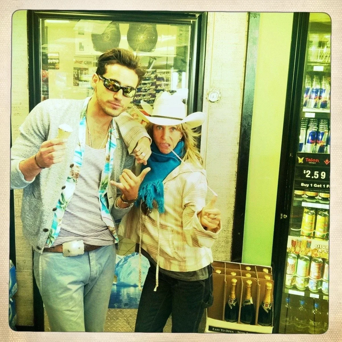 Two people posing inside a convenience store; a man with sunglasses and a casual jacket holding an ice cream, and a woman wearing a cowboy hat, a blue scarf, and a striped jacket making a playful gesture.