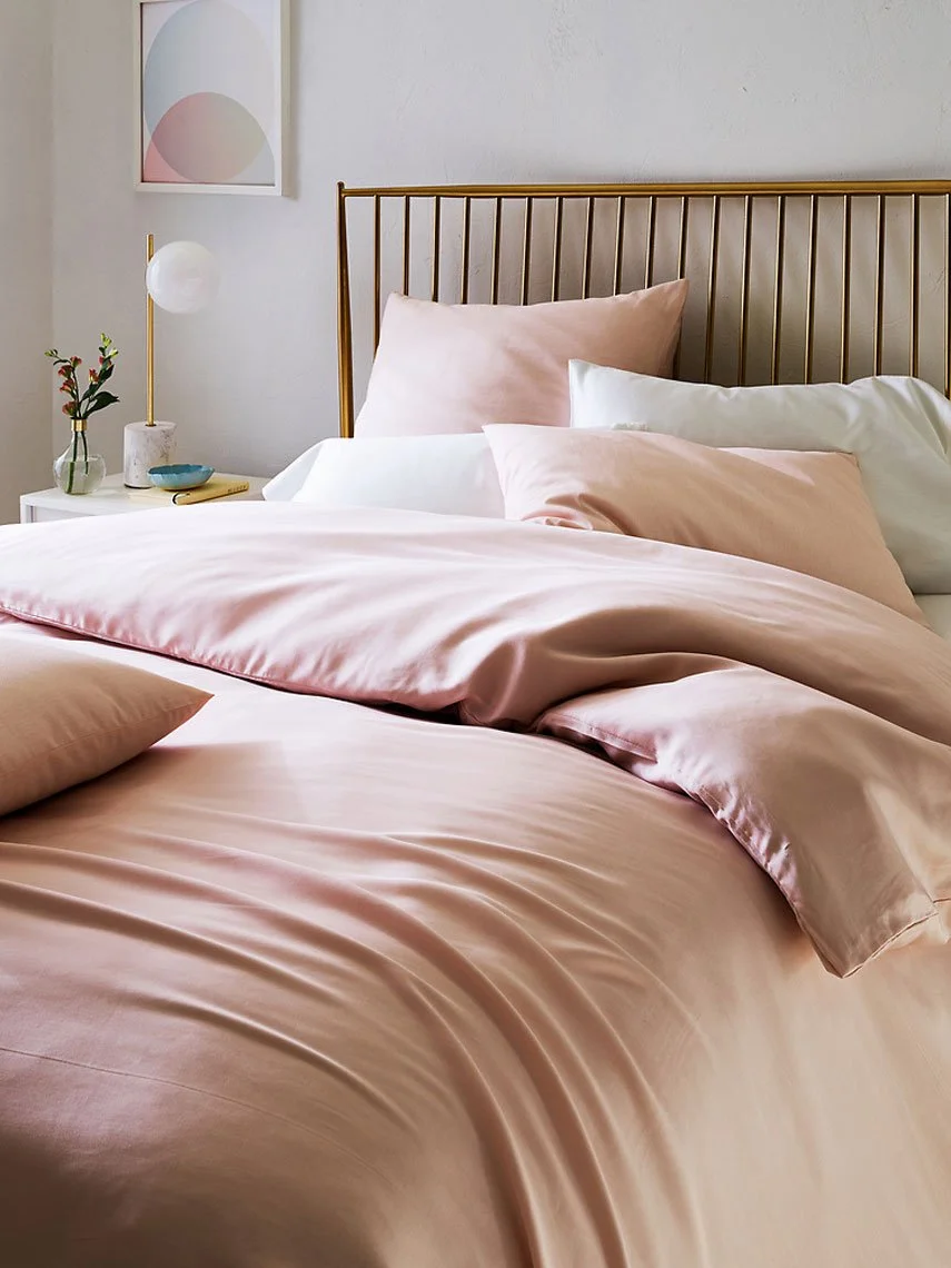 A neatly made bed with pink satin sheets and pillows in a bedroom with minimalist decor, including a bedside table with a small vase of flowers and a modern lamp.