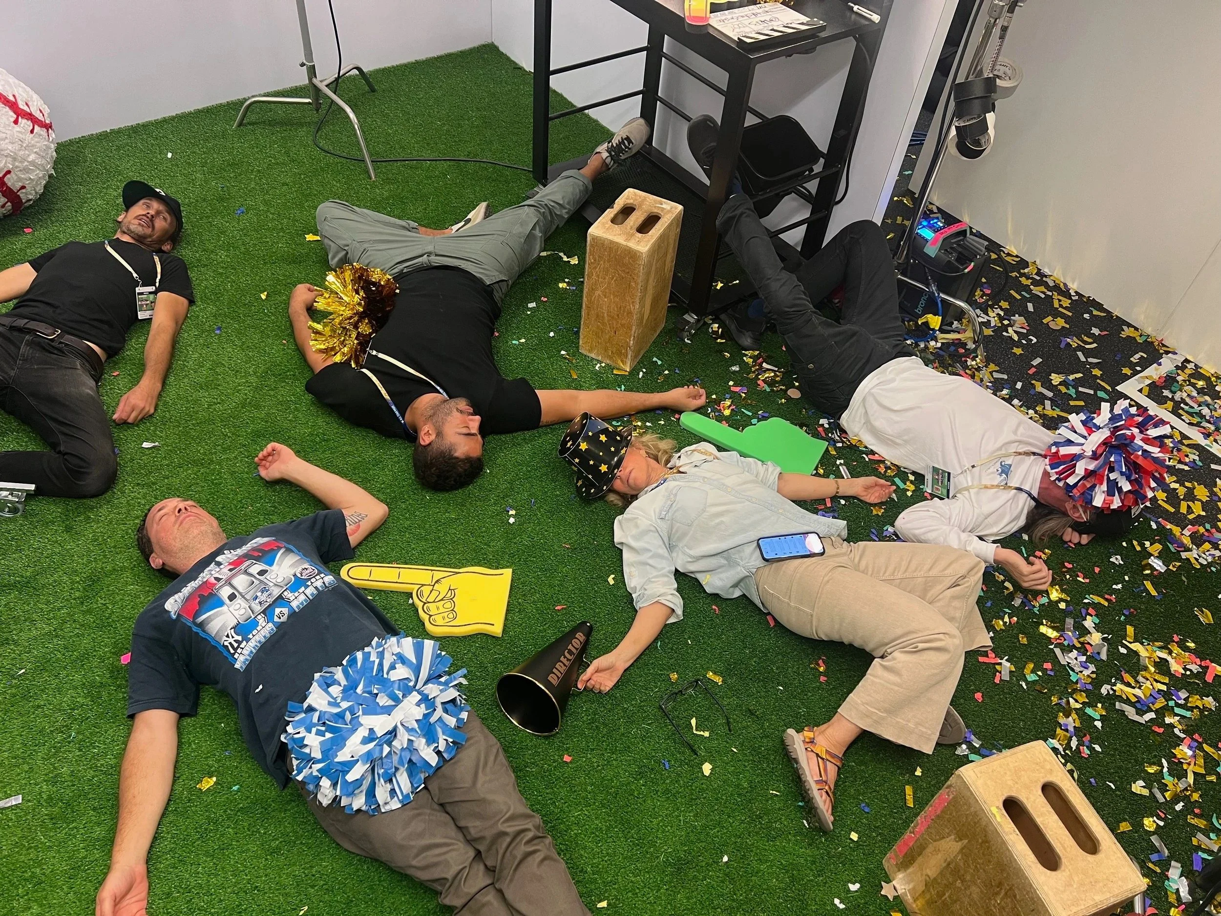 People lying on the floor amidst party decorations and confetti, with some wearing party hats and pom-poms, in an indoor setting.