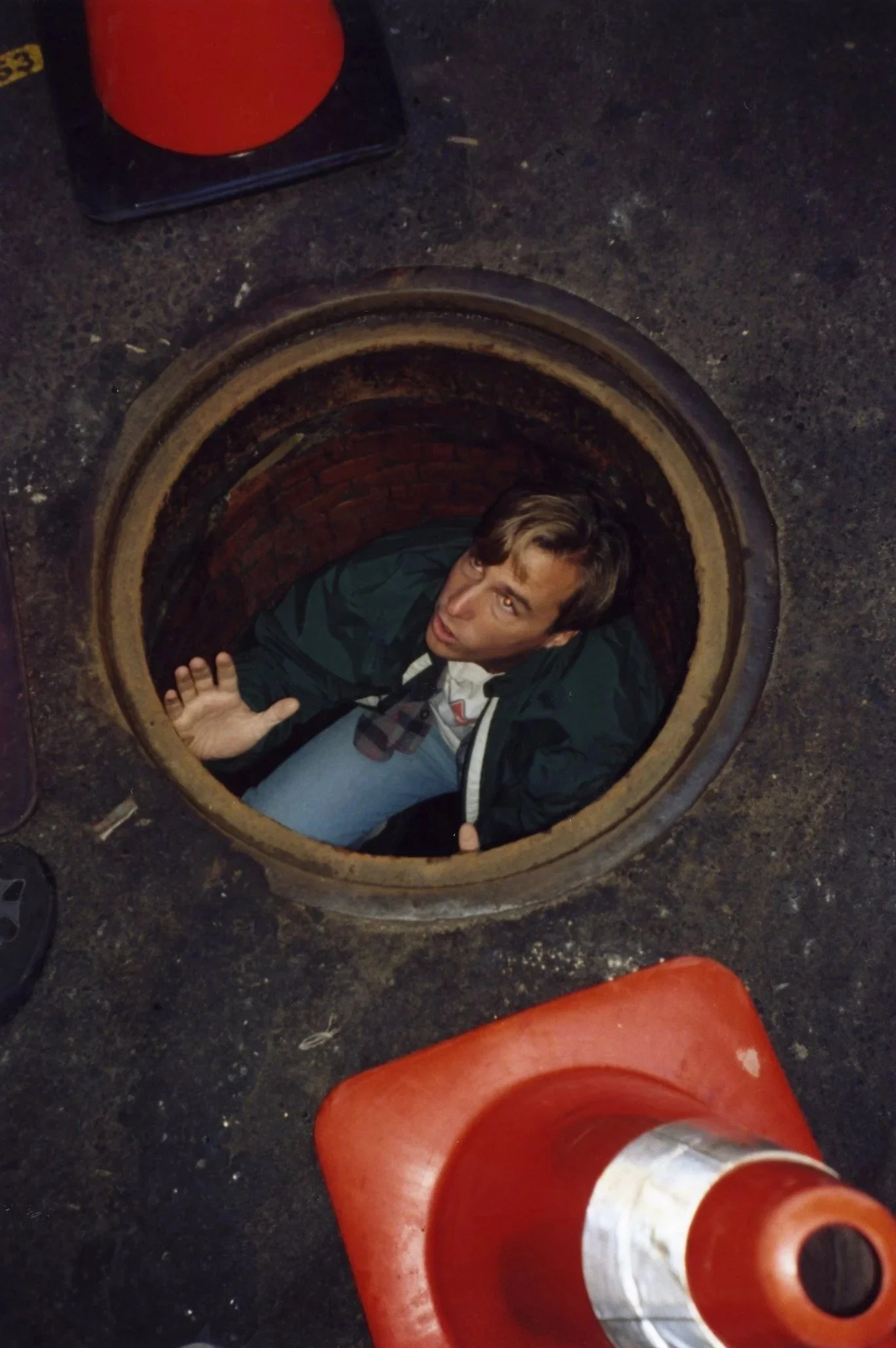 A man is falling into an open manhole on a paved street, with orange traffic cones surrounding the opening and a red object nearby.