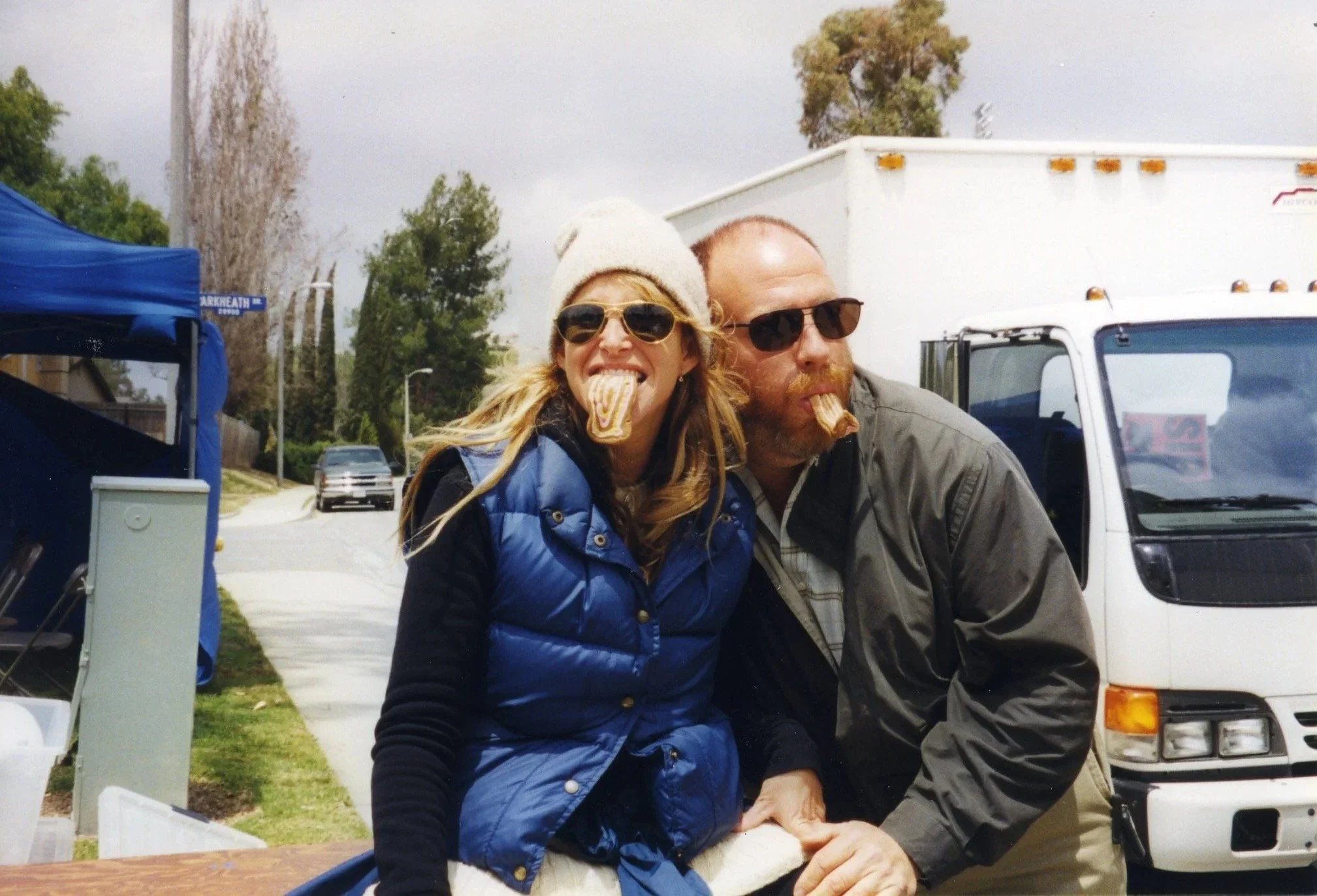 A woman and a man with beards and sunglasses are sitting outdoors at a table, eating what appears to be pastries, each holding a pastry in their mouths. They are smiling and enjoying a casual moment.