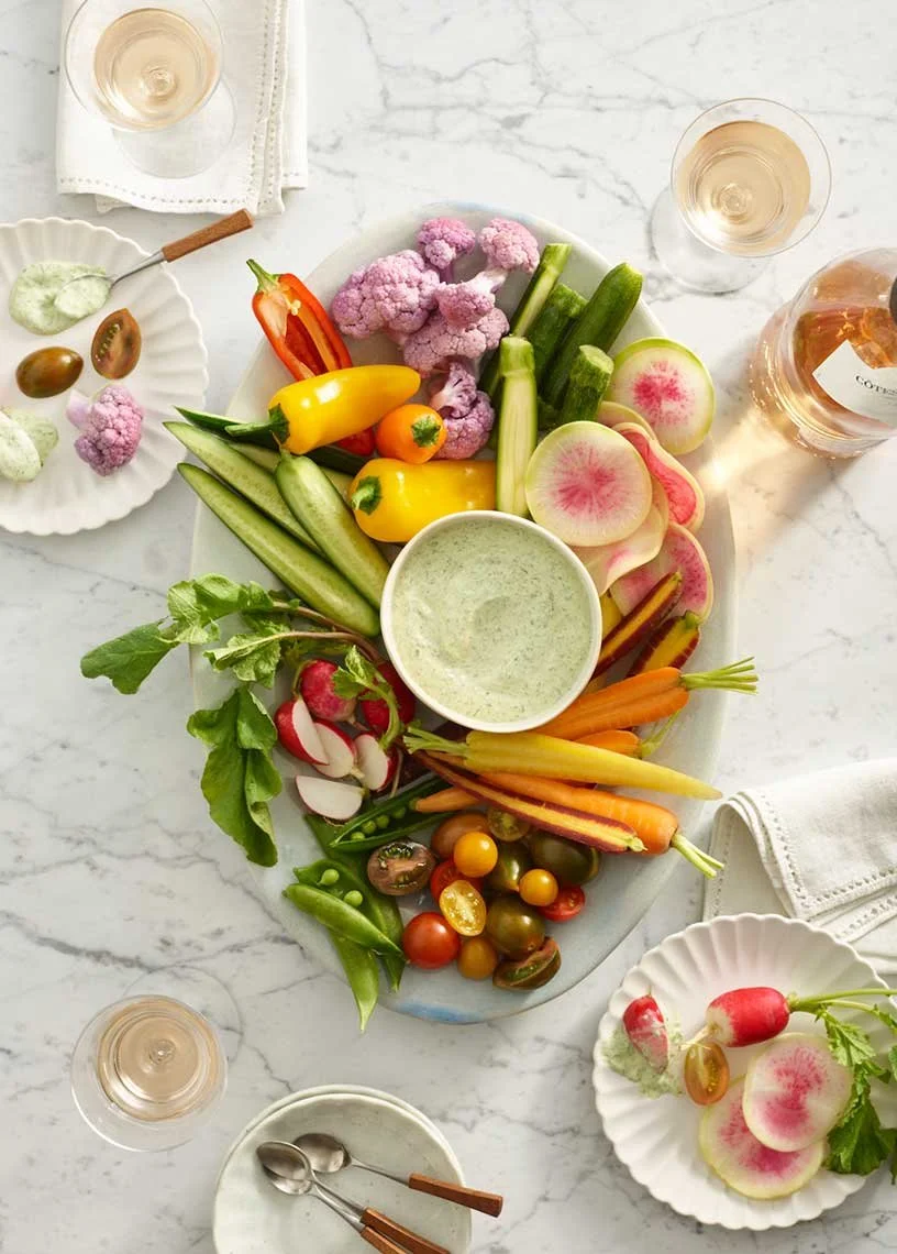 A platter of fresh vegetables including cherry tomatoes, cucumbers, radishes, carrots, cauliflower, and yellow and red peppers, with a dipping sauce in the center, surrounded by glasses of white wine and small plates with radishes and vegetables.