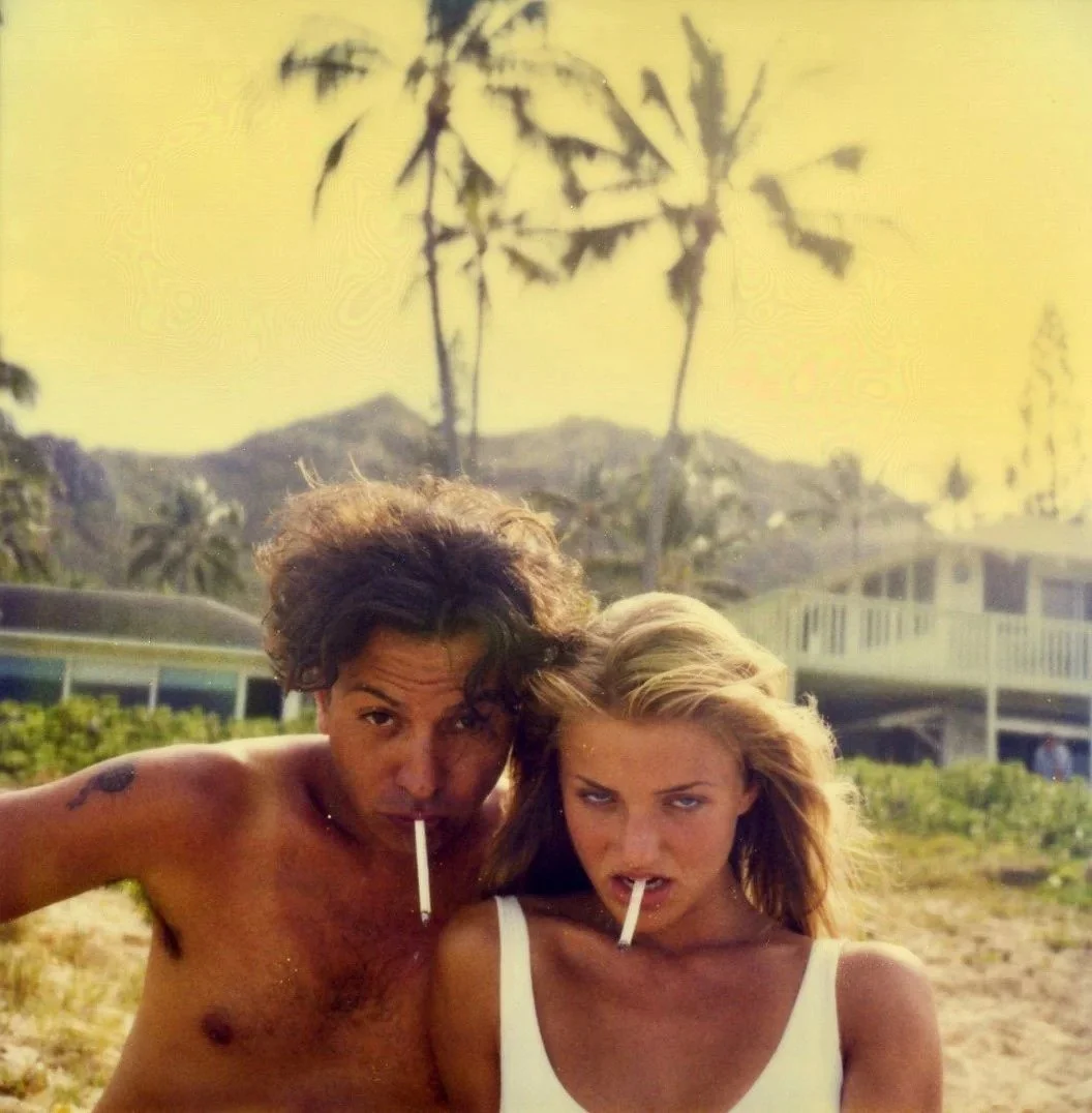 A shirtless man and a woman in a white tank top with cigarettes in their mouths, standing closely outdoors with palm trees and a house in the background.