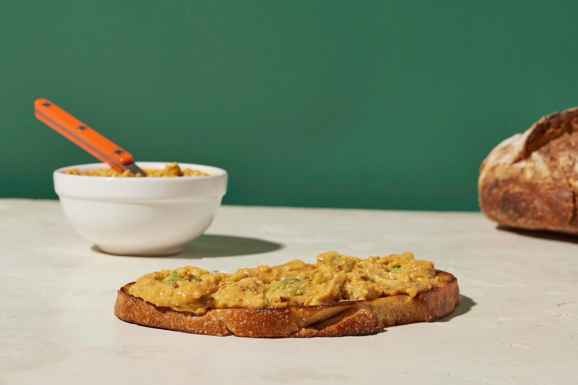 A slice of toasted bread topped with guacamole, placed on a white surface. In the background, there is a bowl of guacamole with a serving spoon inside, and a loaf of bread partially visible on the right side. The background is green.