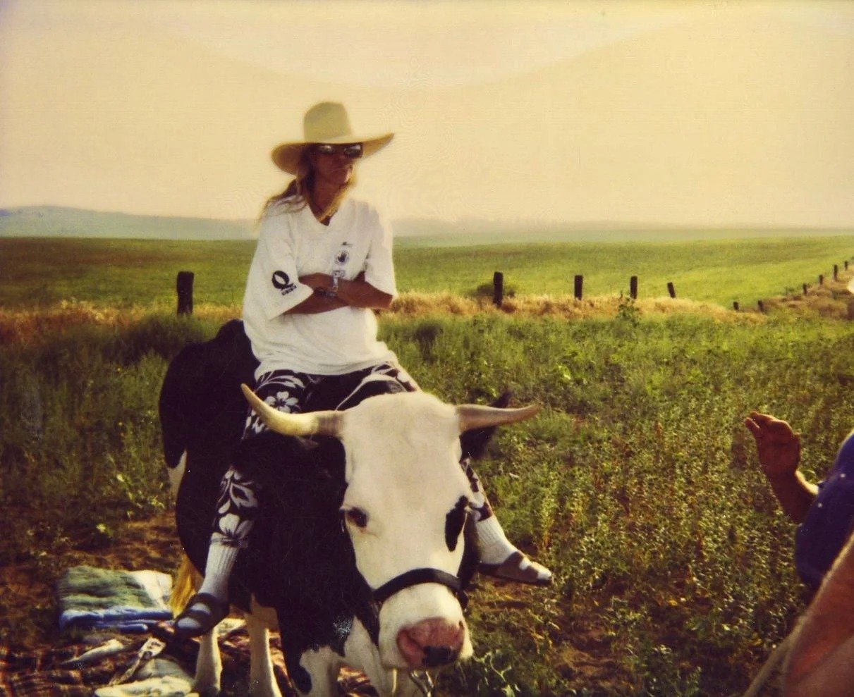 A woman wearing a large sun hat, sunglasses, and a white shirt is sitting on a black and white cow in a green field.