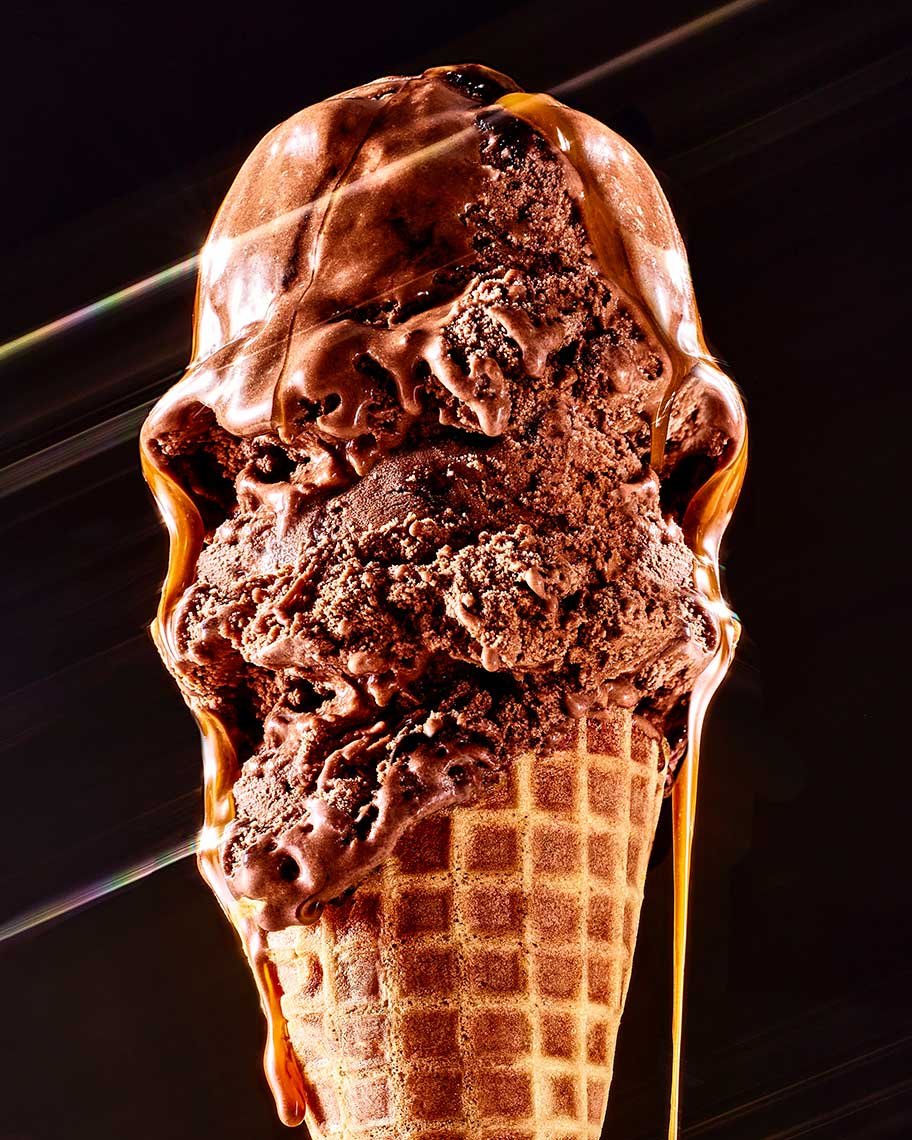 Close-up of a triple-scoop chocolate ice cream cone with melted chocolate dripping down, against a dark background.