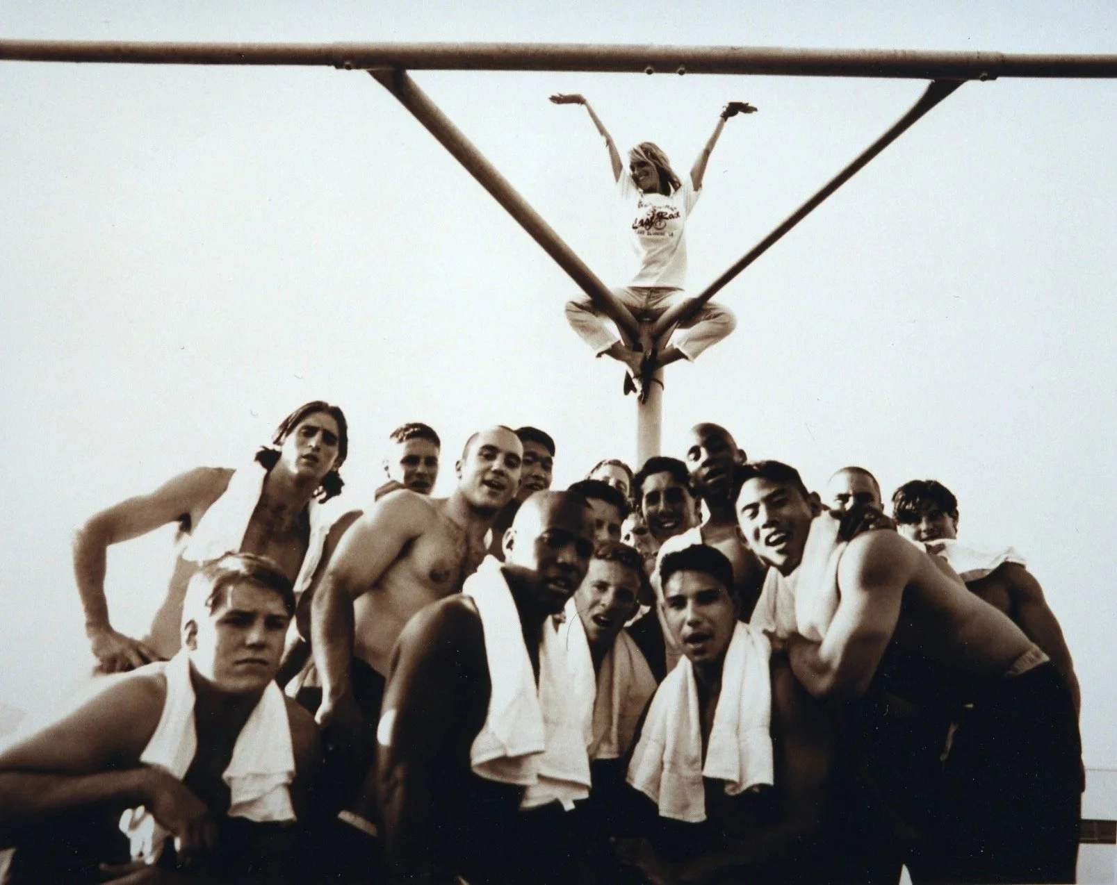 Black and white photo of a group of shirtless men with towels around their shoulders, posing together with a woman sitting on a pole above them, raising her arms in the air.