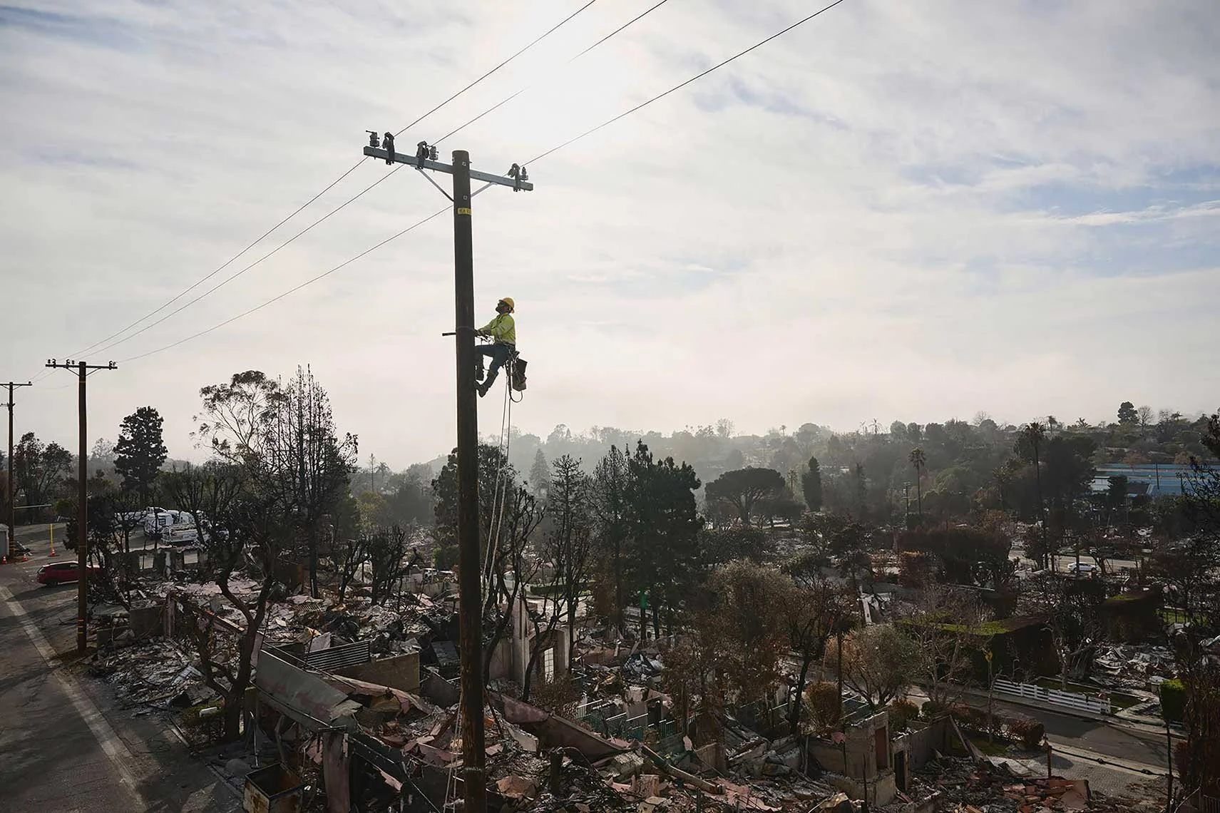 A worker in safety gear installing or repairing power lines on a utility pole during daytime, with a background of trees and damaged structures indicating a recent disaster.