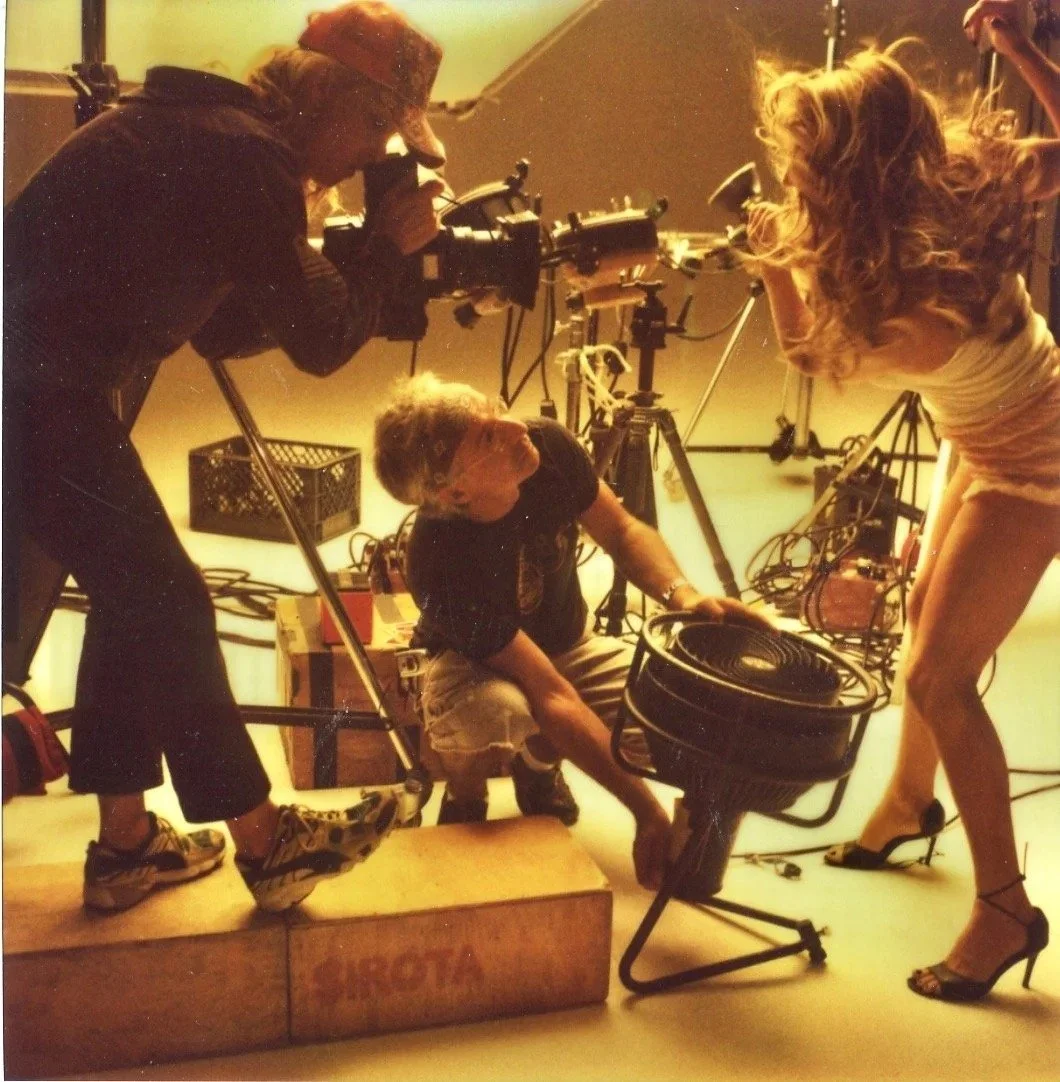 Behind-the-scenes of a film set with a camera operator, a woman in high heels, and a man adjusting lighting equipment, amid various filming gear.