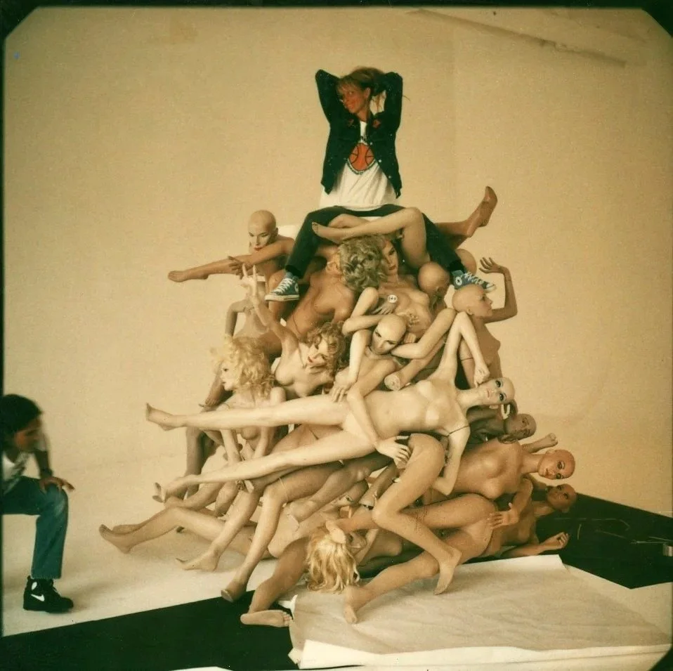 An art installation featuring a pile of mannequin parts and dolls with a girl sitting on top, and a person sitting to the side.
