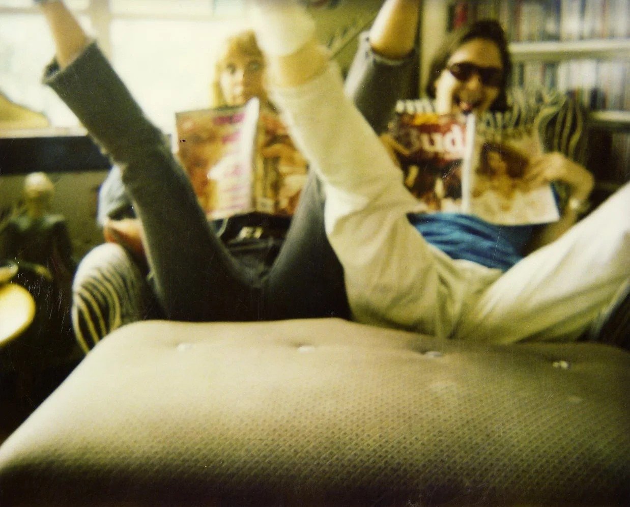 Two people lying on a sofa in a library, eating snacks, with one wearing sunglasses.