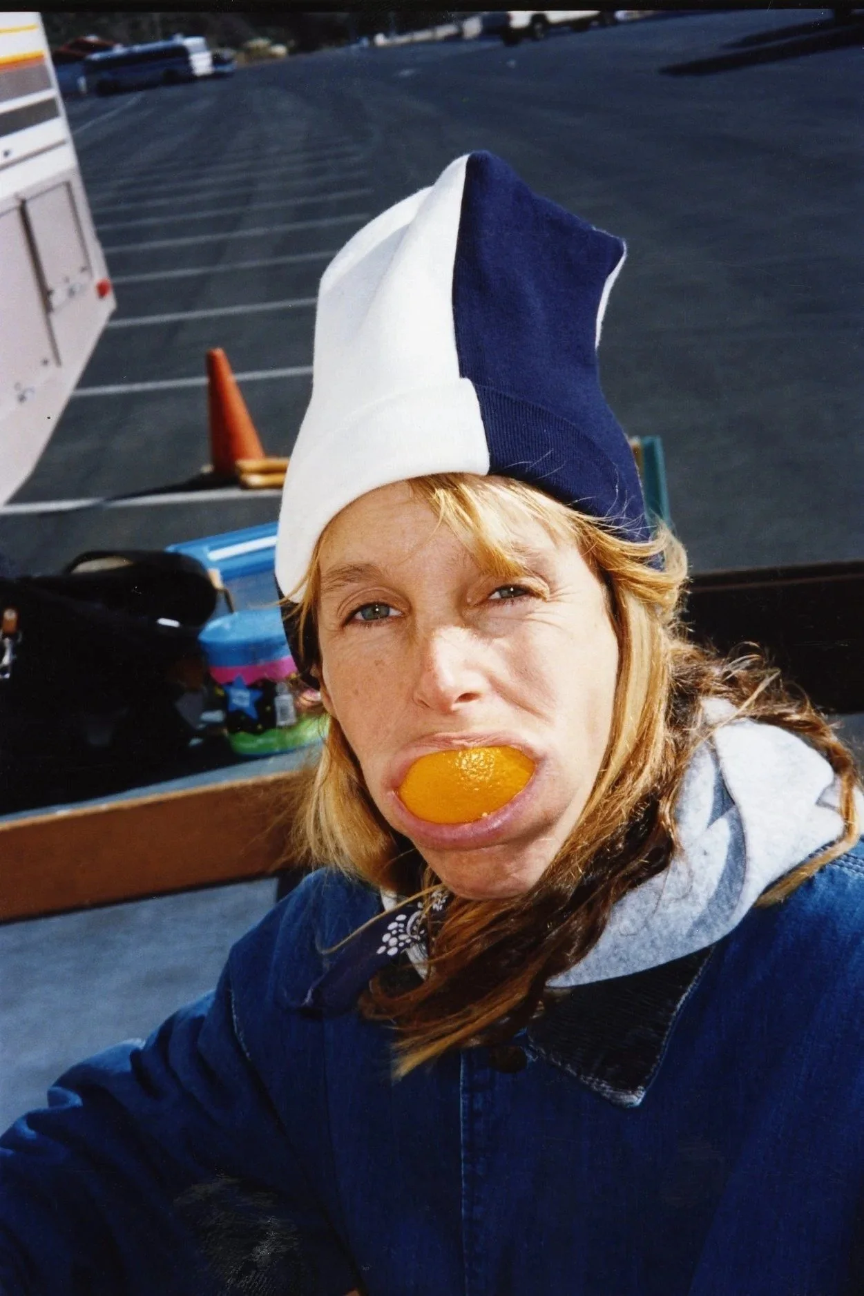 Woman with red hair and freckles wearing a blue and white hat, holding an orange in her mouth, sitting outdoors in a parking lot with orange cone and equipment in the background.