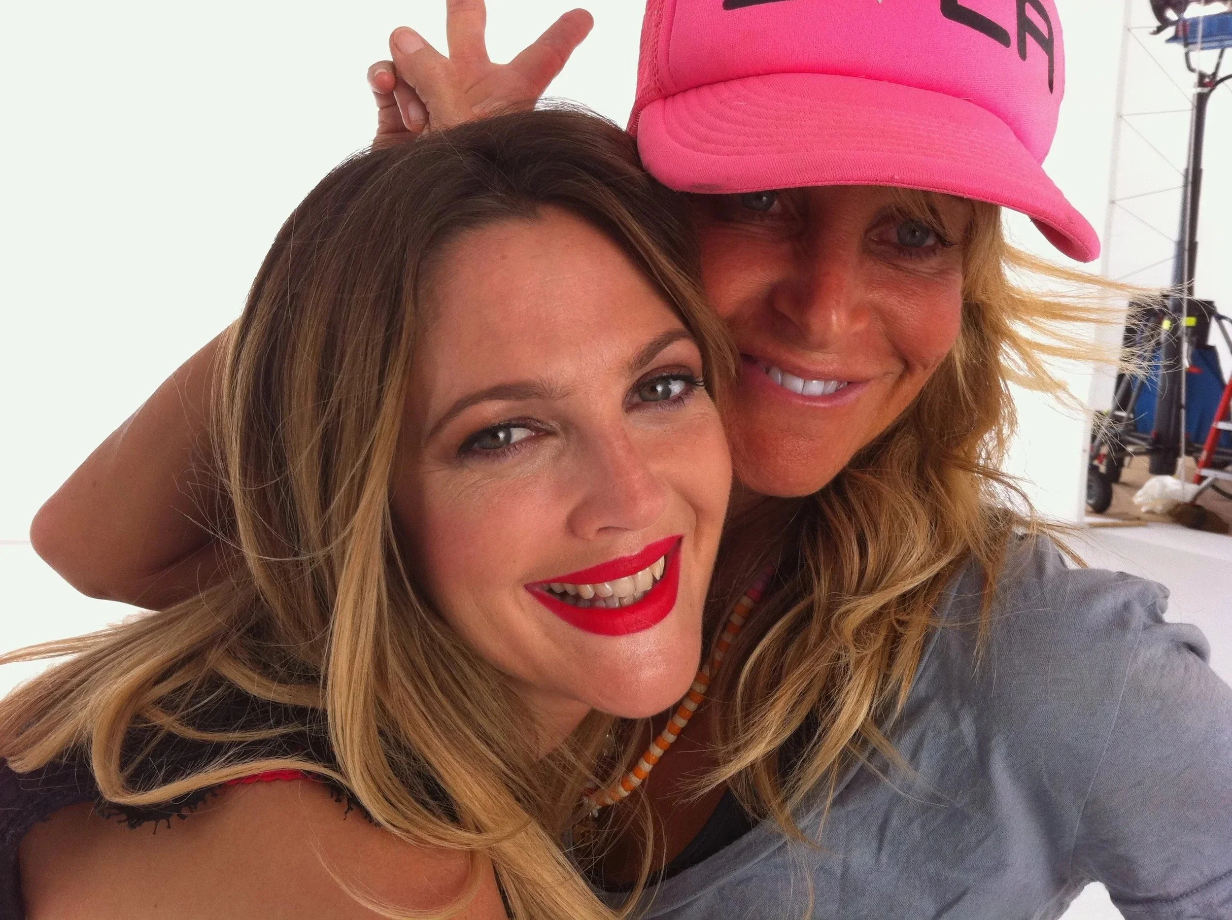 Close-up of two women smiling and posing for a photo, with one wearing a pink cap and the other showing blonde hair and bright red lipstick.