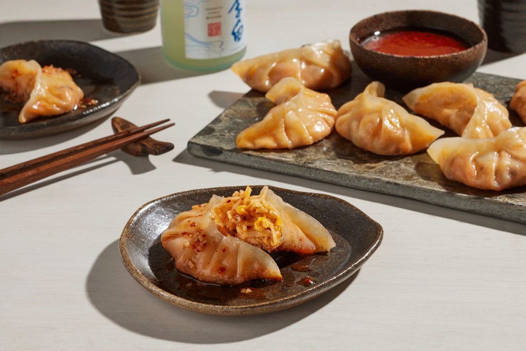 Close-up of steamed dumplings on a ceramic plate and a wooden serving board, with dipping sauces and chopsticks on a white table.