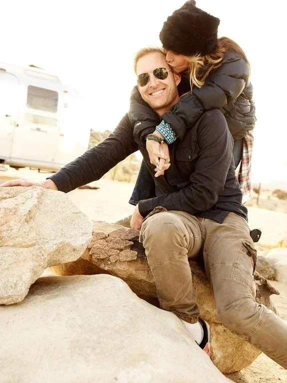 A man with sunglasses smiling while sitting on a large rock, with a young girl hugging him from behind, in a desert setting with a trailer in the background.