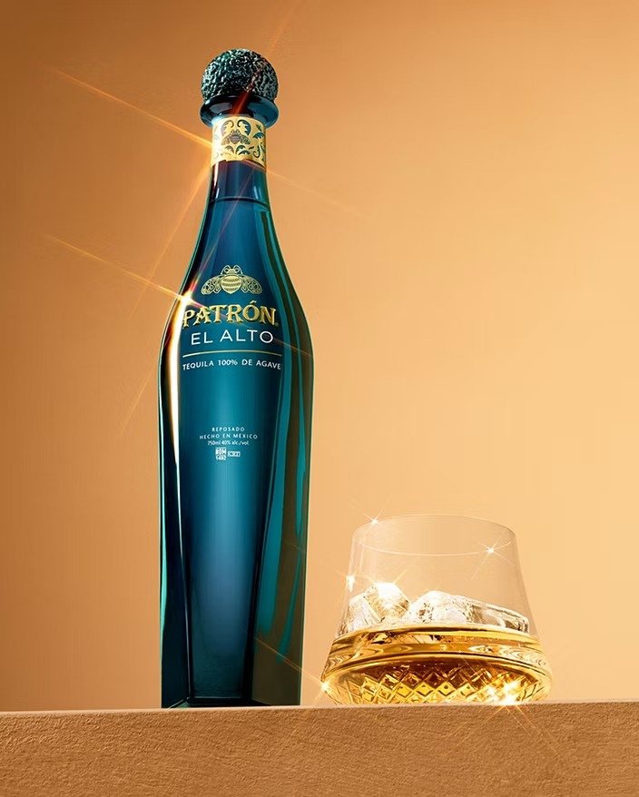 A tall bottle of Patrón El Alto tequila with a black and gold cap, placed on a surface next to a glass of tequila with ice, against a warm, orange background.