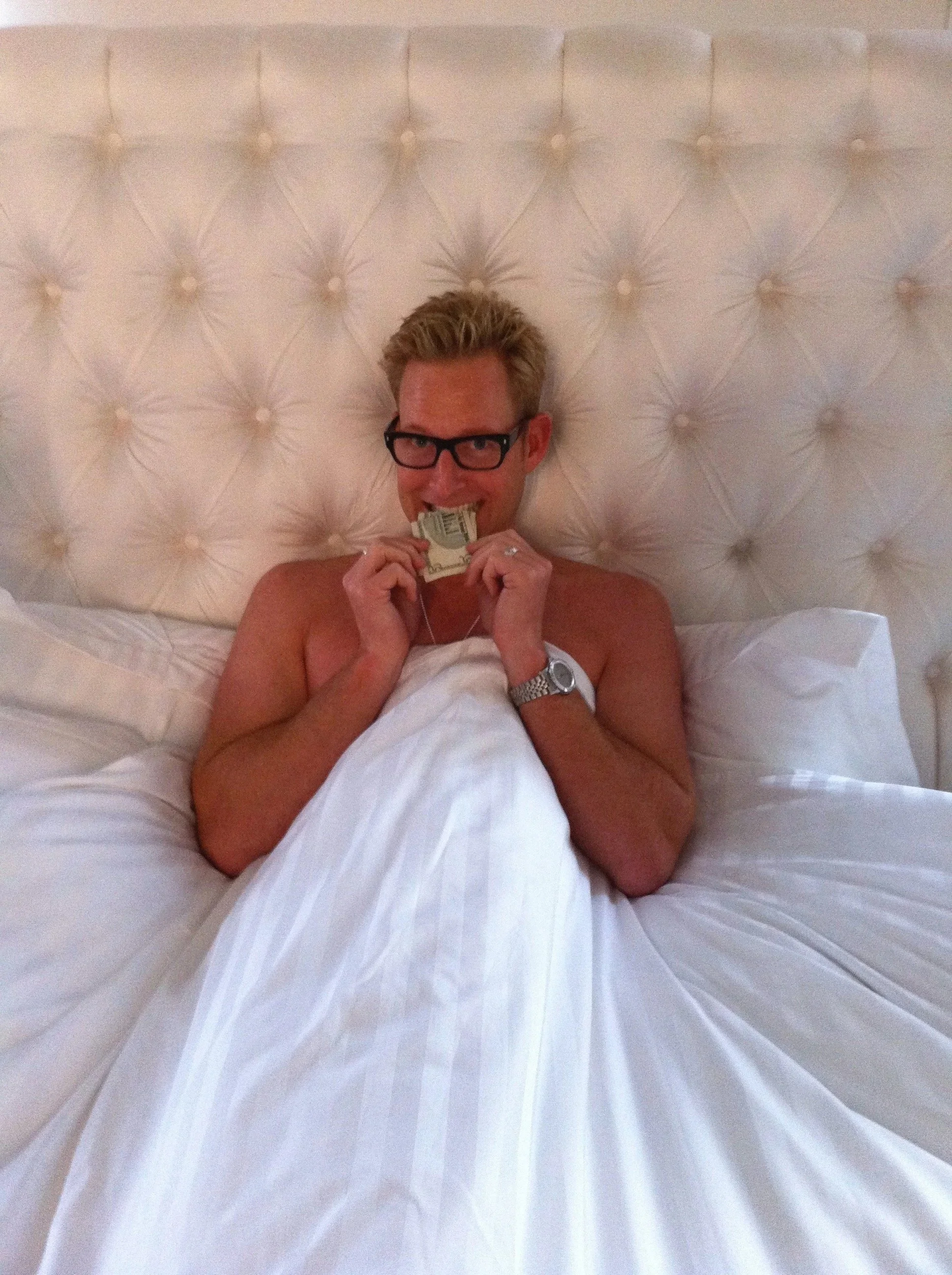A man lying in bed with a white headboard, holding a dollar bill in his mouth, wearing glasses and a wristwatch.