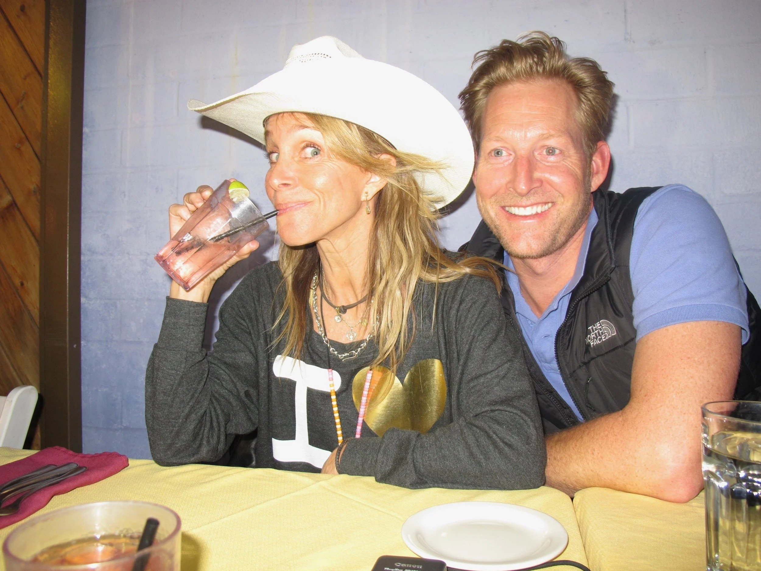 A woman wearing a large white sunhat and a dark shirt drinks a pink beverage with a lime wedge during a meal at a restaurant. A man next to her with light brown hair, a blue shirt, and a black vest smiles at the camera. The table has a yellow tablecl
