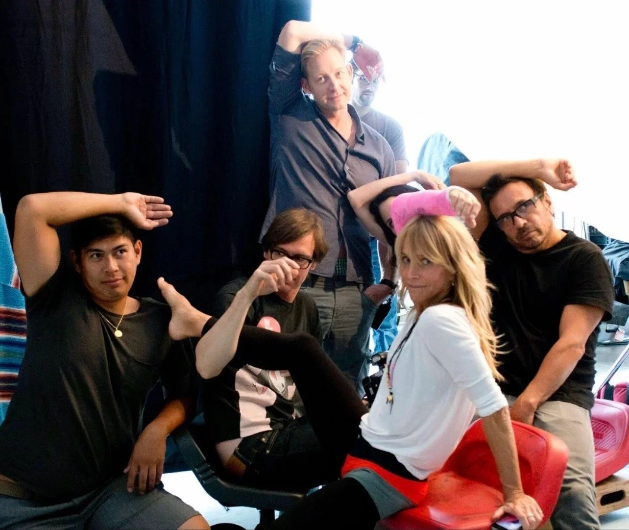 Group of five people posing in a playful manner indoors. Two are sitting, one standing in the background, and two posing with arms bent. The woman in white is sitting on a red chair, and others are making humorous gestures.