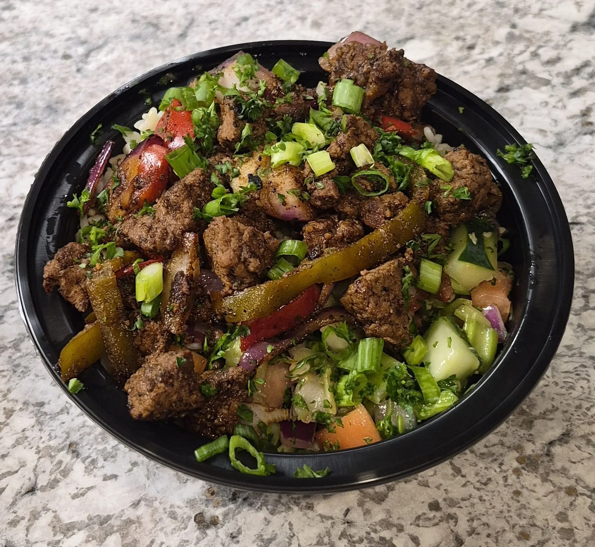 A black plastic bowl filled with cooked meat, vegetables, and chopped green onions sits on a speckled gray countertop.