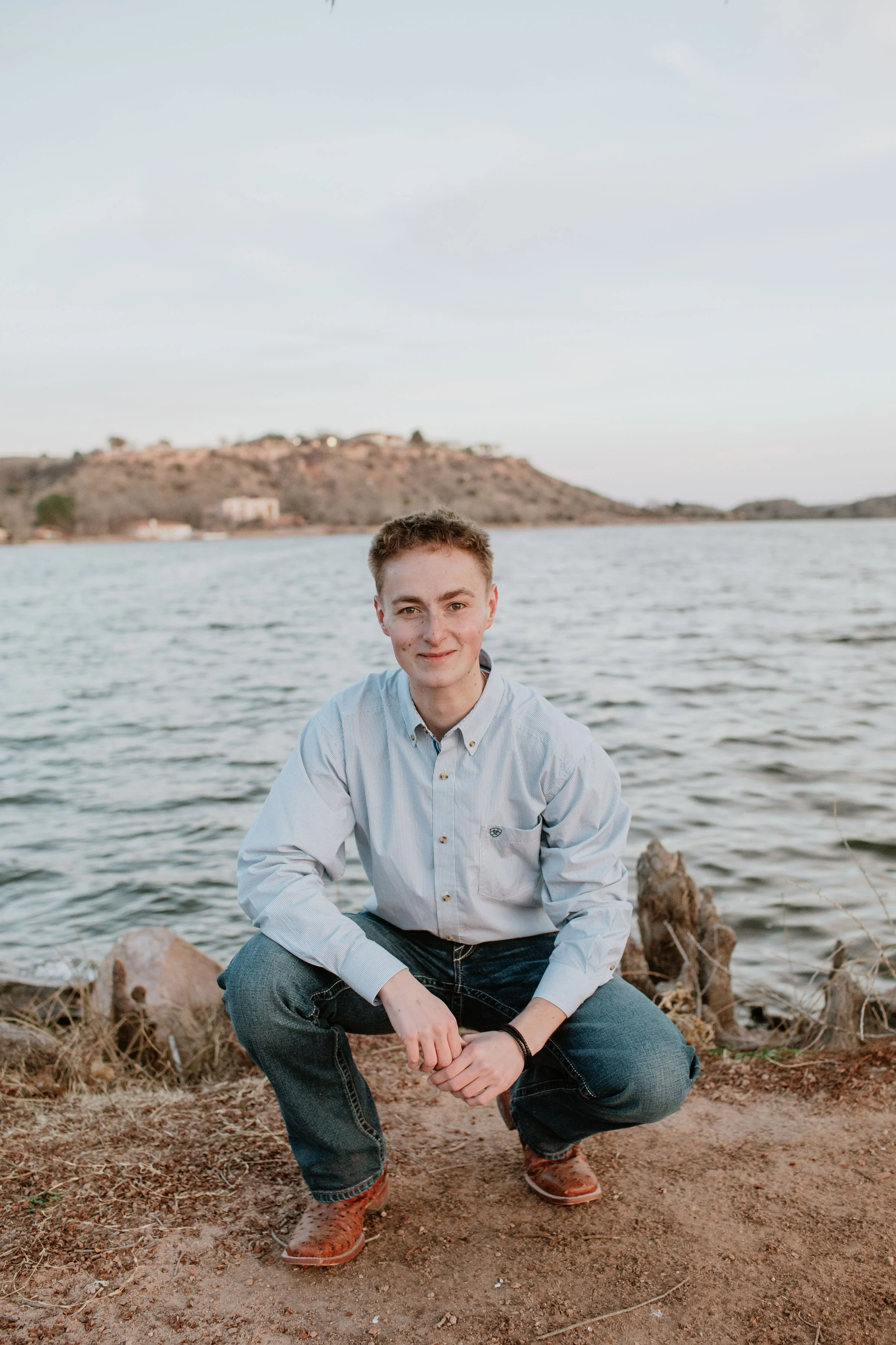 A young man with light skin, short light brown hair, and blue eyes, wearing a light blue button-up shirt, black jeans, and brown shoes, is crouching on a rocky shoreline near a body of water, with a hill and some buildings in the background.