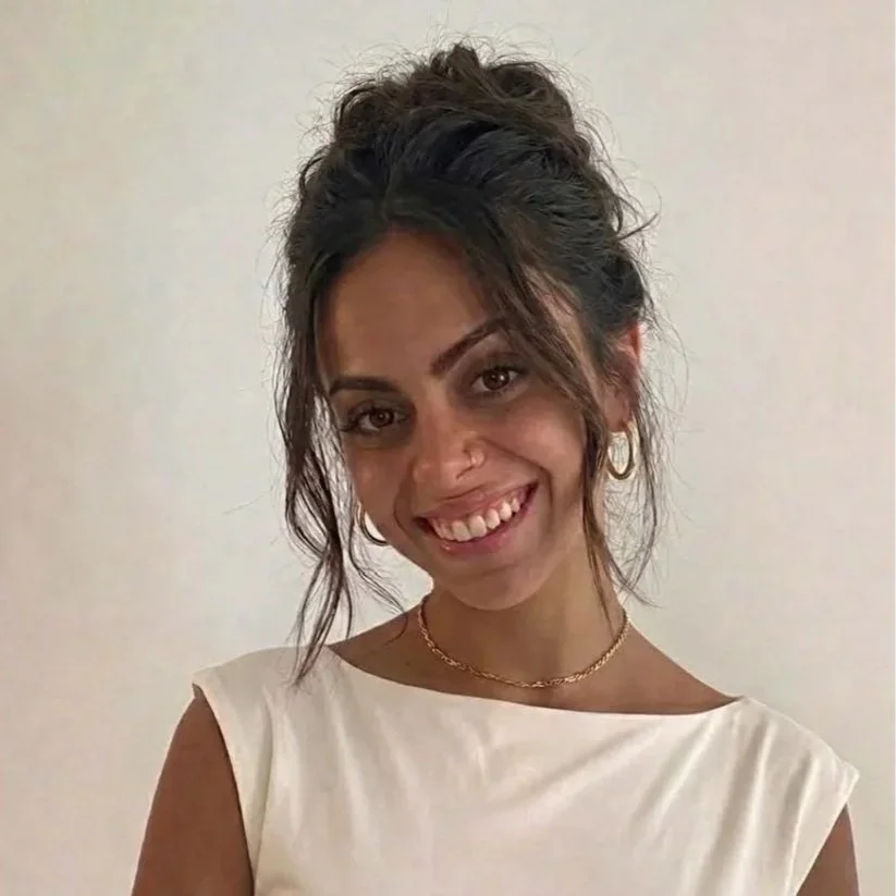 A young woman with curly dark hair tied up, wearing a white sleeveless top, gold hoop earrings, a gold necklace, and smiling at the camera.
