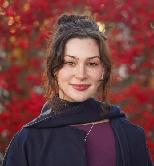 A young woman with wavy dark hair tied in a bun, smiling, wearing a dark jacket and a purple top, standing outdoors with red fall leaves background.