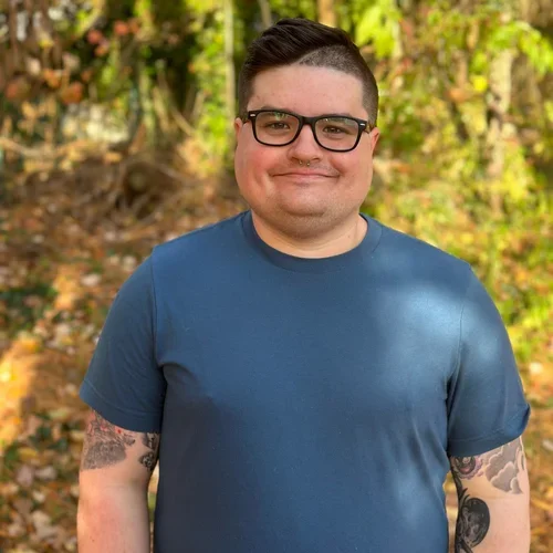 Young man smiling outdoors with trees and fall leaves in the background, wearing a navy blue t-shirt and glasses.