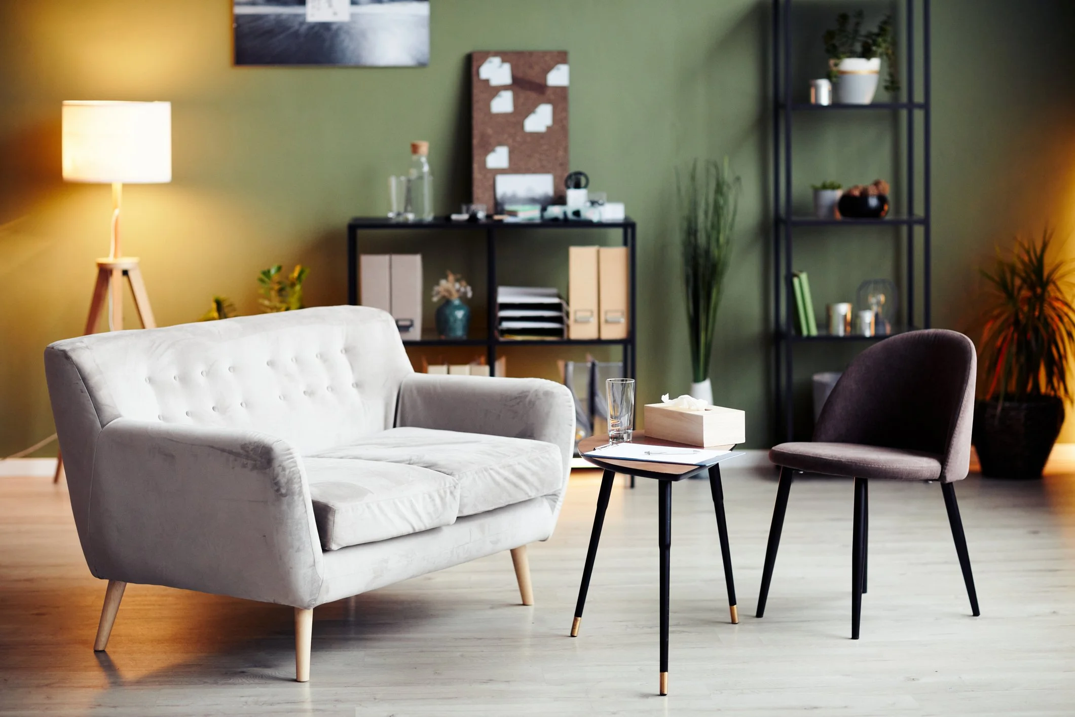 A cozy living room with a white tufted armchair, a small black side table with a glass of water, and a brown chair nearby. In the background, a black shelving unit holds plants and decorative items, and a green wall features wall art and a bulletin board. A floor lamp provides warm lighting.